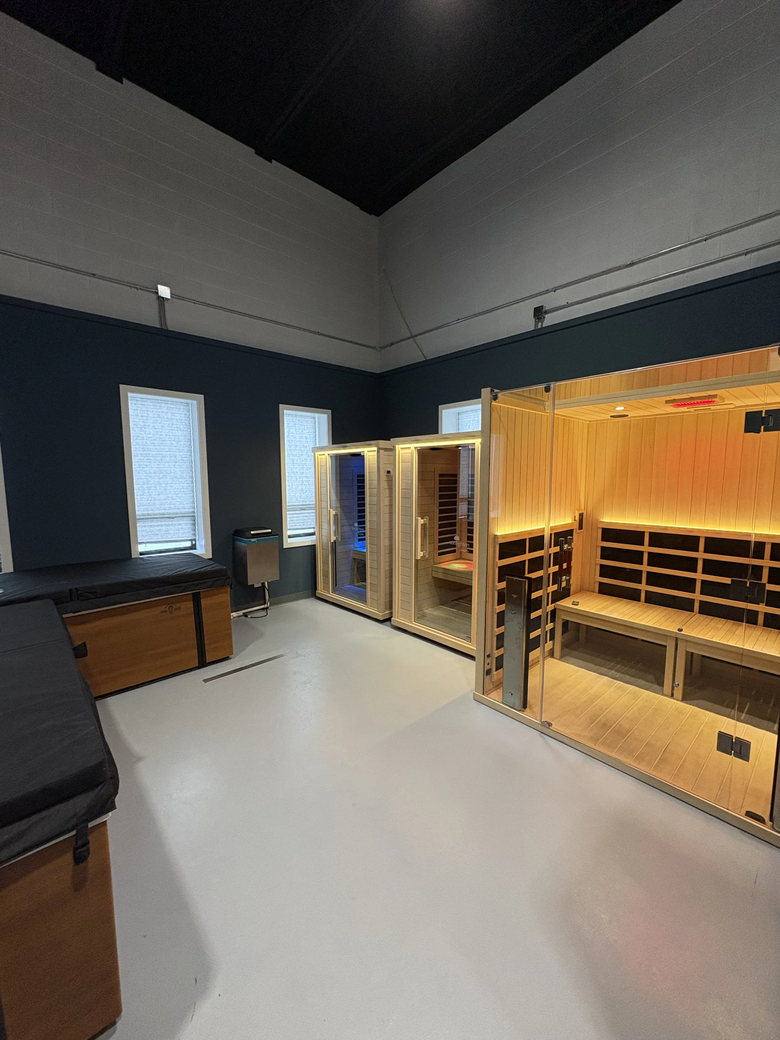 Interior of a room with three wood and glass infrared sauna units, blue walls, three small windows, gray floor, and some furniture.