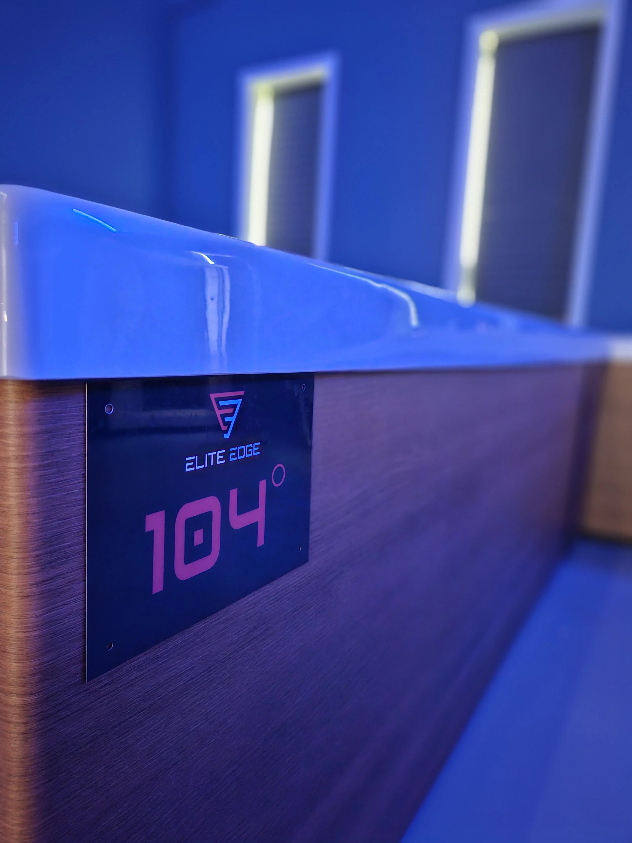 Close-up of a temperature sign on a hot tub, showing 104 degrees Fahrenheit in red text, with a blue-lit countertop and two vents on a blue wall in the background.