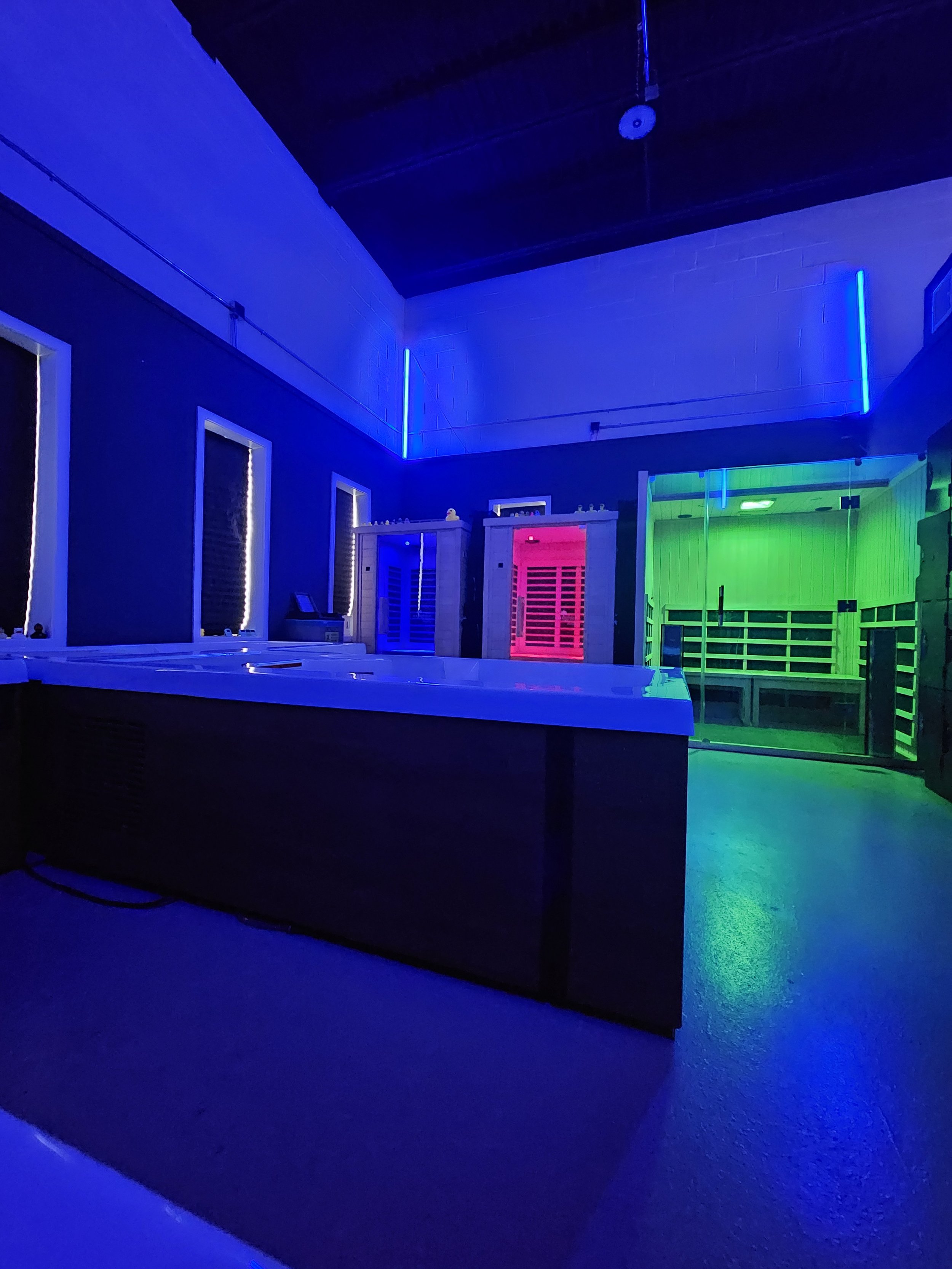 A room illuminated with colorful LED lights, featuring blue, red, and green lighting accents on the walls and ceiling. There are windows with frames, some objects on a surface, and a visible door or partition.