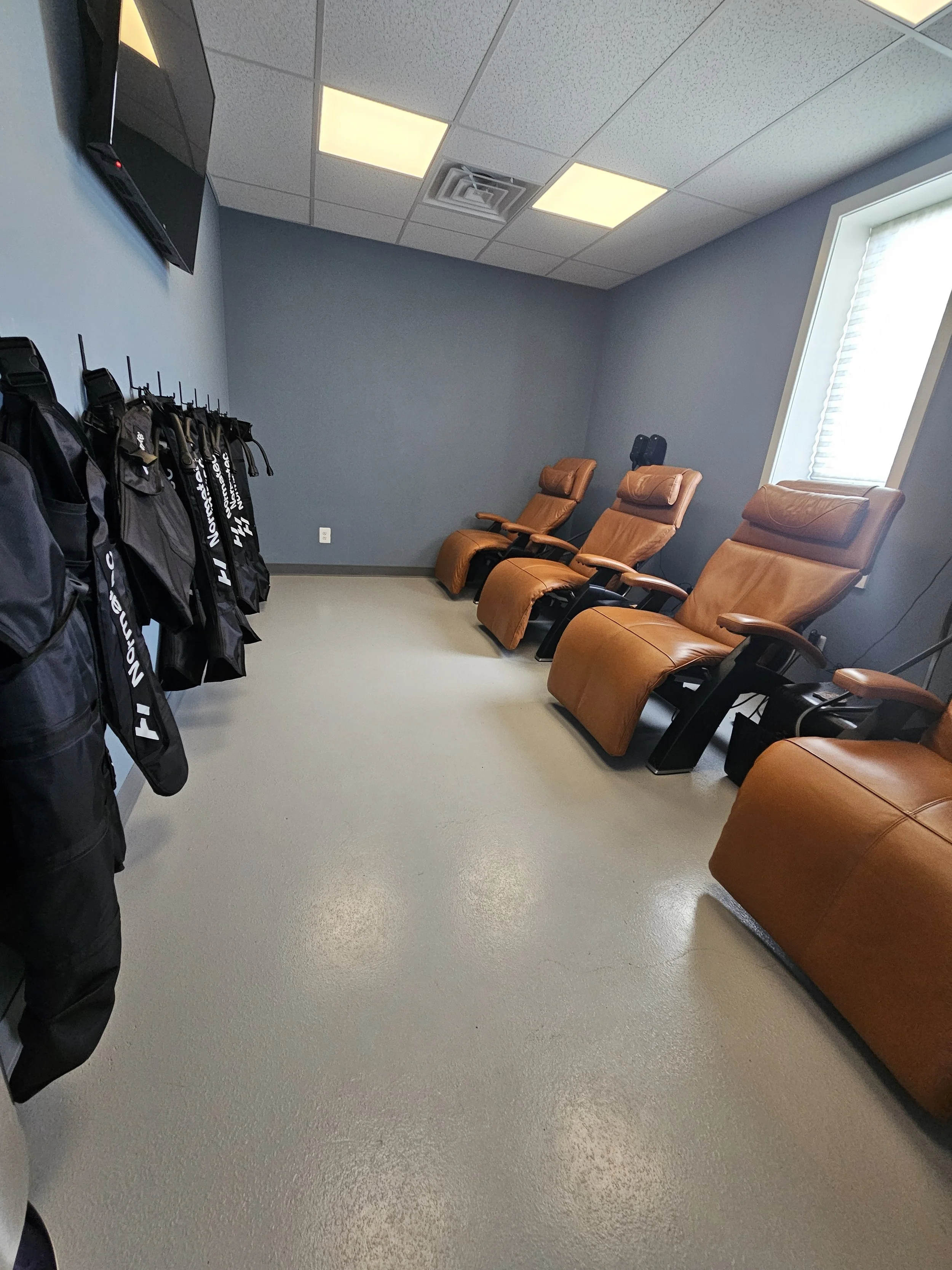 A room with three brown massage chairs lined up against a wall near a window, and a row of black jackets hanging on hooks on the opposite wall.