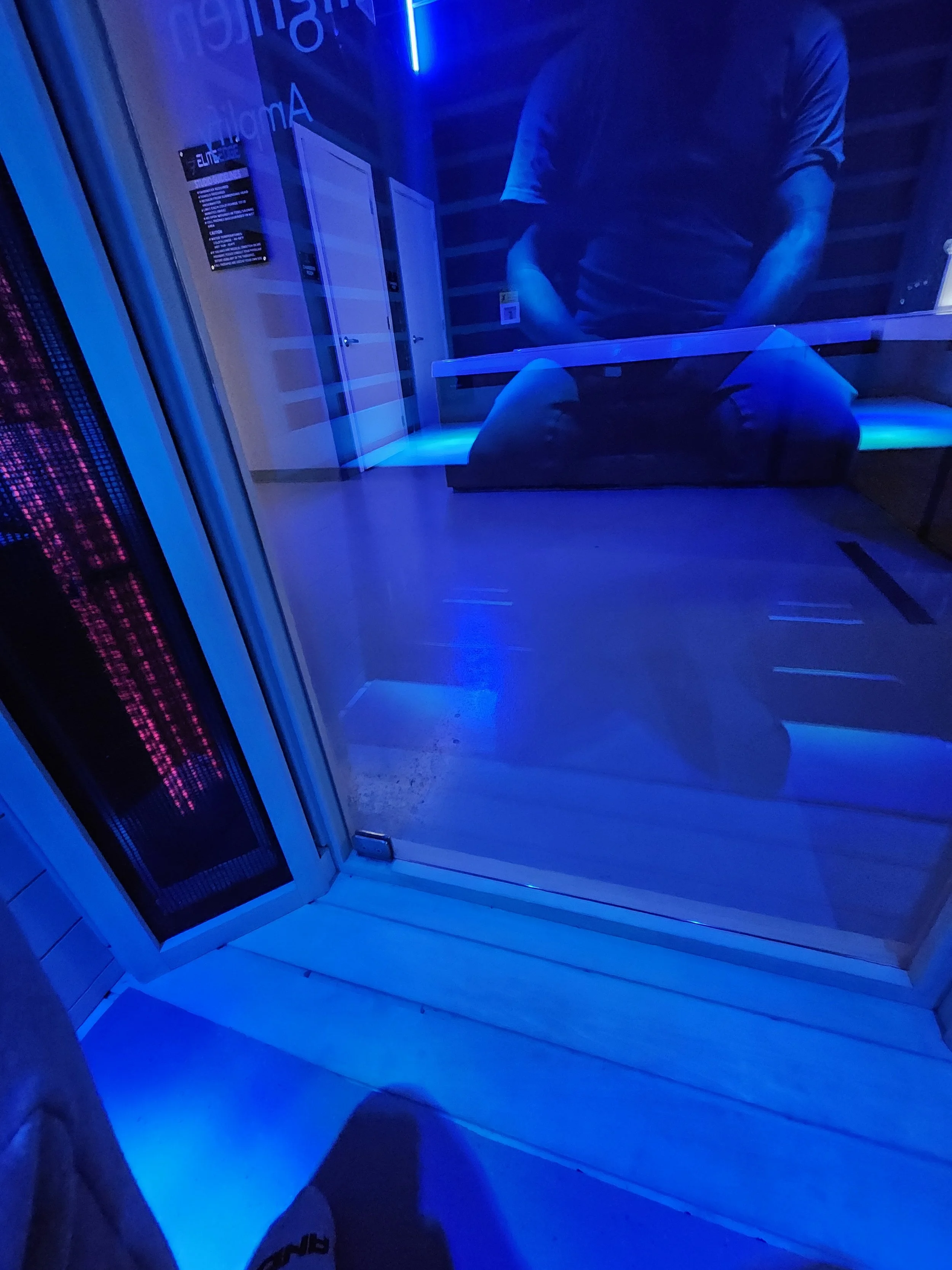 Reflection of a person in a glass door with blue and pink neon lighting, and a person sitting on a bench inside a modern room.