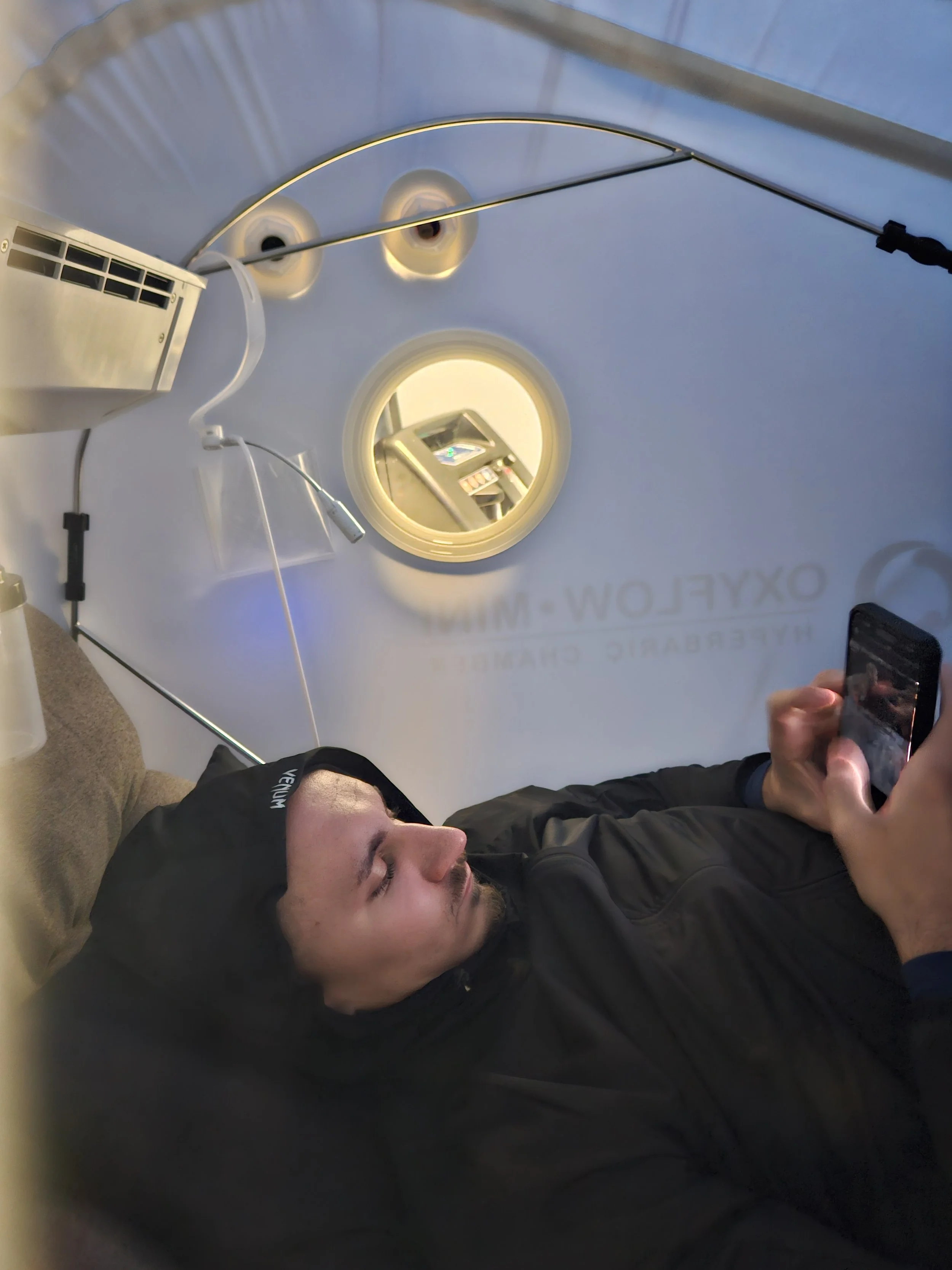 A man lying down inside a hyperbaric chamber, looking at his phone.