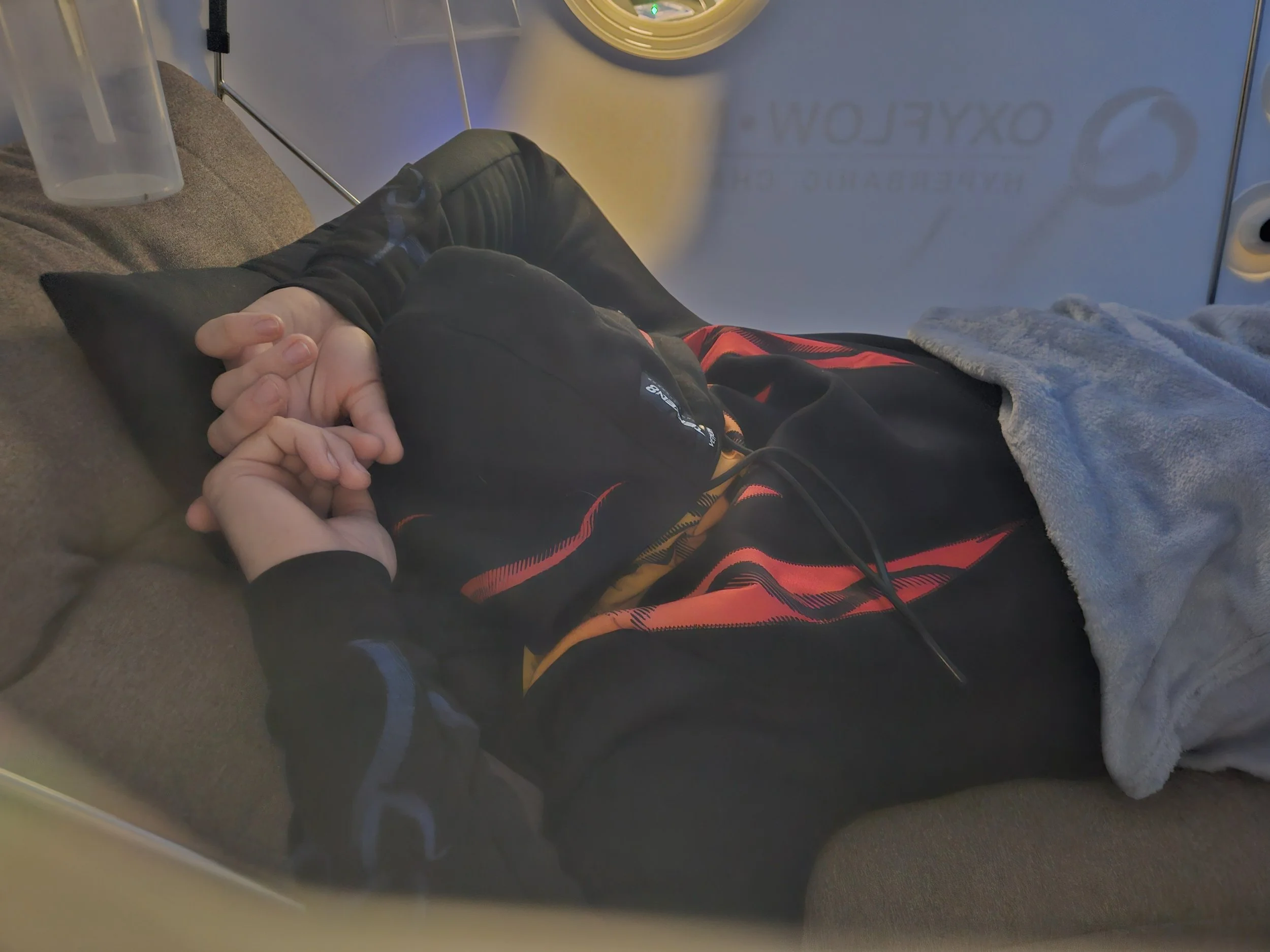 Person lying on a motorized vehicle or amusement ride, holding hands with another person, with a water cup nearby and a sign in the background.