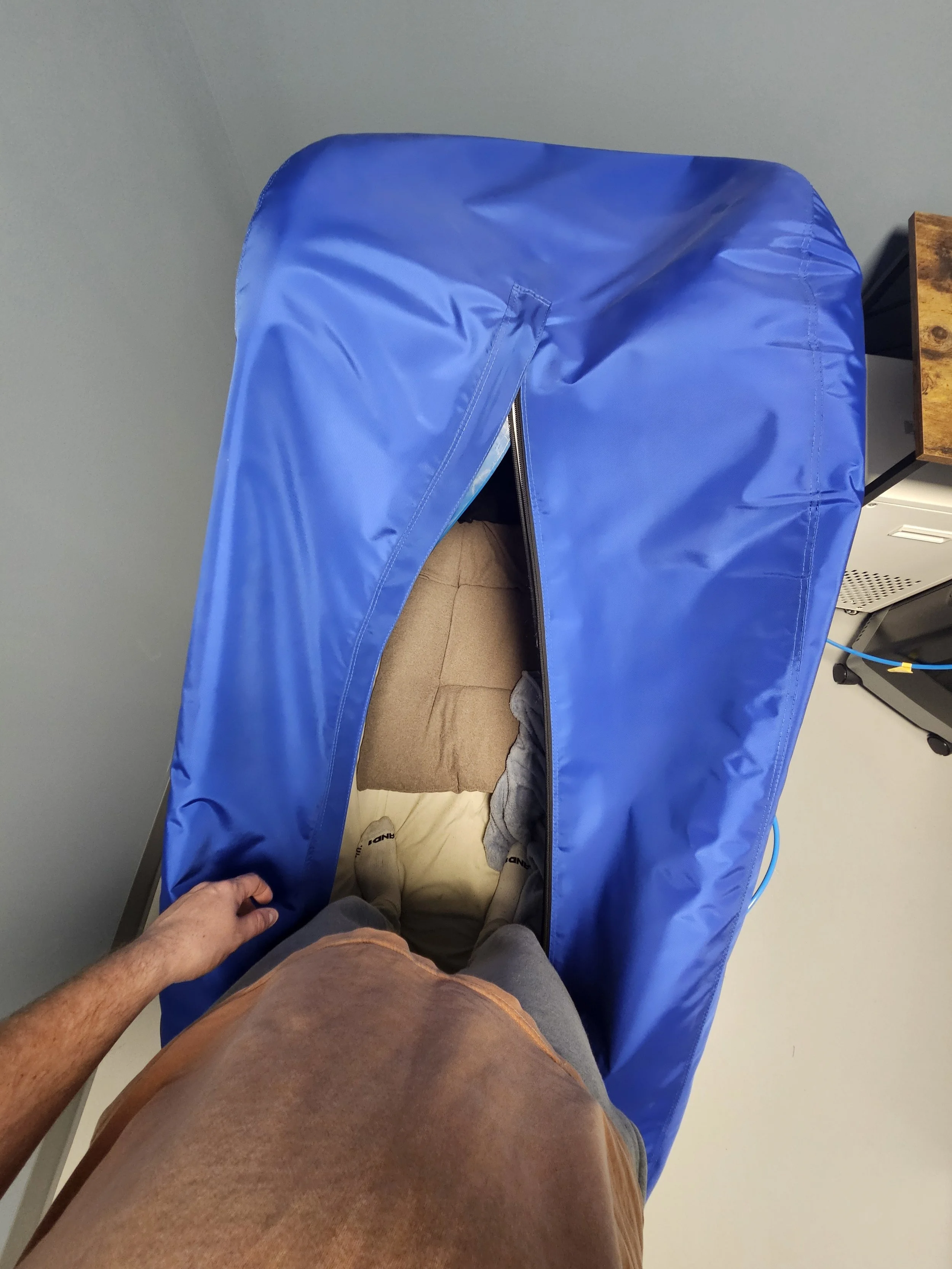 Hand holding the edge of a Hyperbaric Oxygen Chamber, with a person wearing a tan shirt and gray shorts standing underneath. The setting appears to be indoors with a gray wall and a wooden table on the right.
