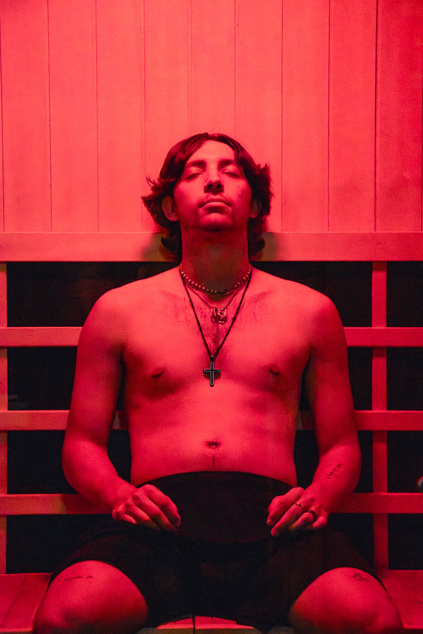A shirtless young man with short dark hair, wearing necklaces including a black cross and a gold pendant, sits with eyes closed in front of a wooden paneled background illuminated with red light.