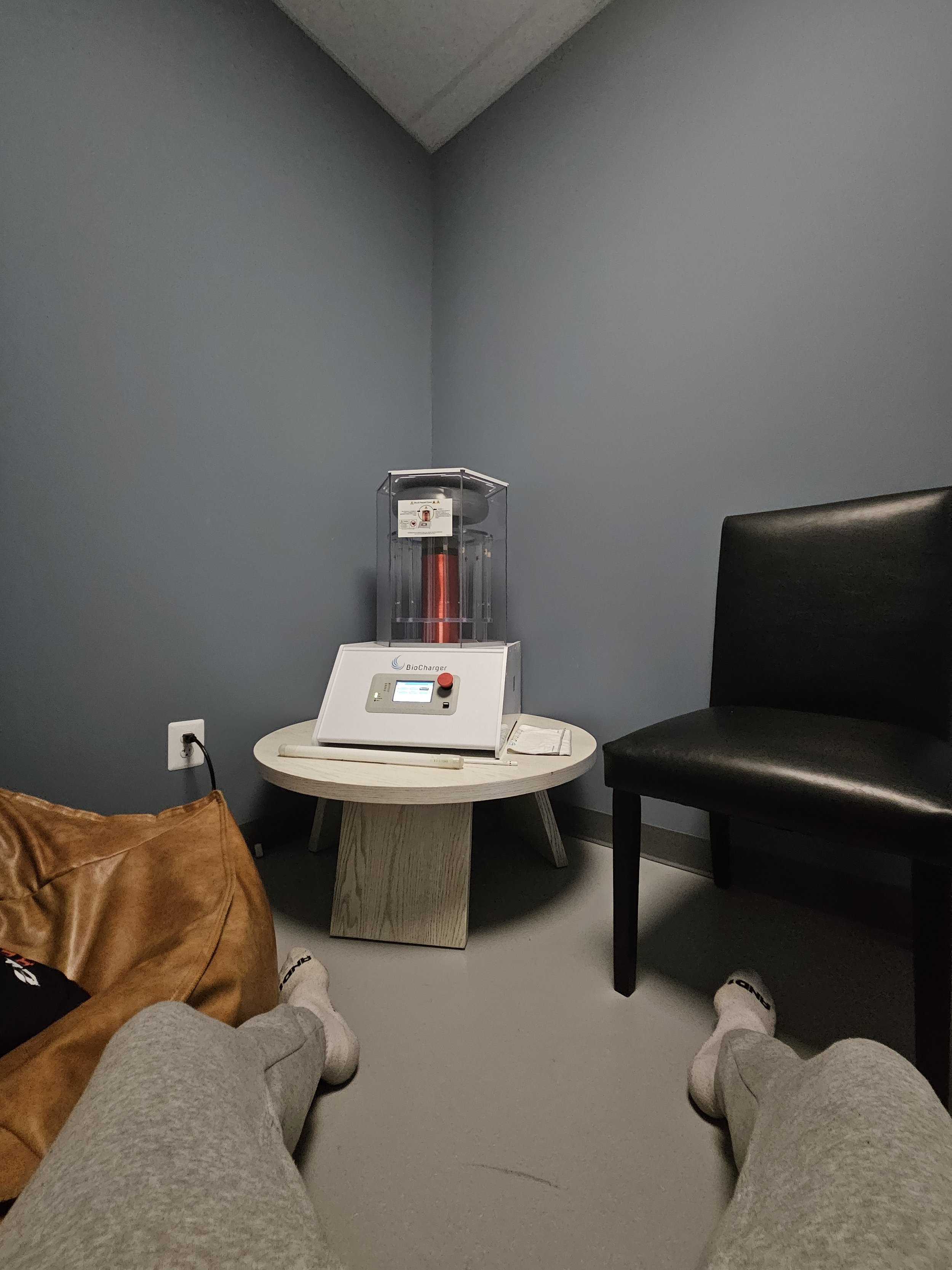 View of a vaccination room with a person's legs and feet in the foreground, a brown bag on the floor, a black chair, and a medical device on a small round table against a blue wall.