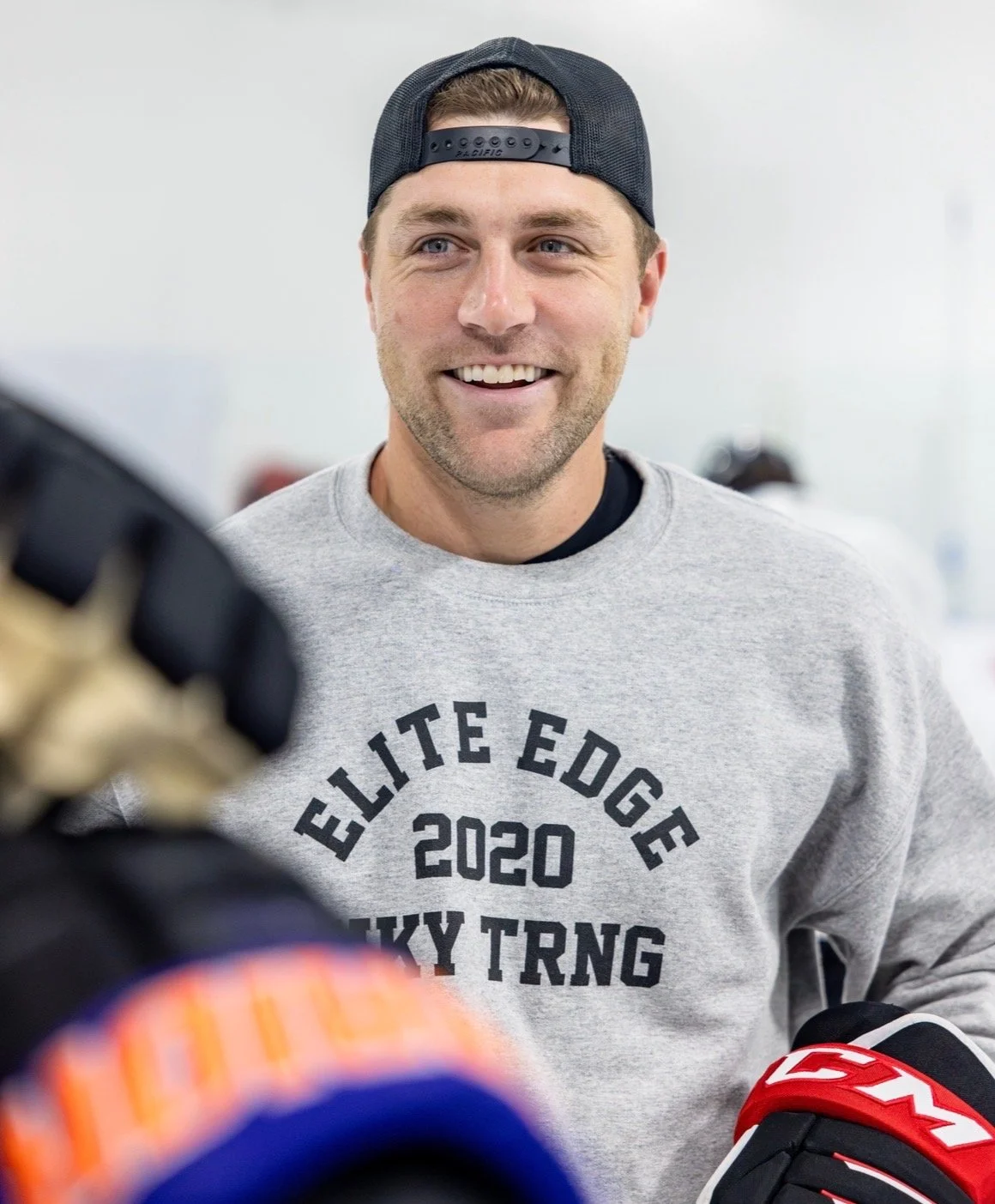 A smiling man wearing a gray sweatshirt with 'Elite Edge 2020 Hockey TRNG' printed on it and a black backwards baseball cap, engaged in a conversation with someone in hockey gloves.