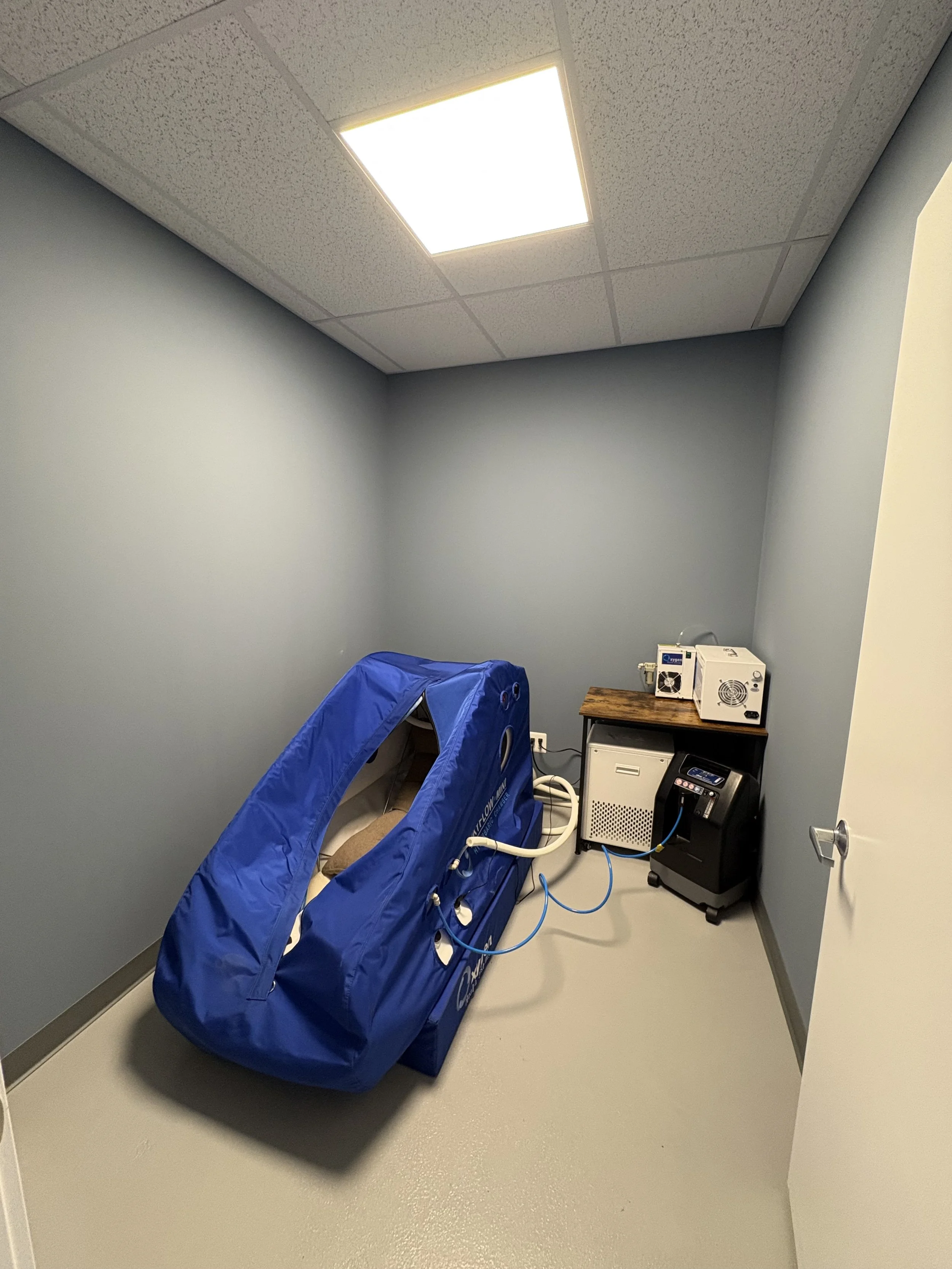 A hyperbaric oxygen therapy chamber in a small room with gray walls, gray flooring, and a ceiling with a fluorescent light panel.