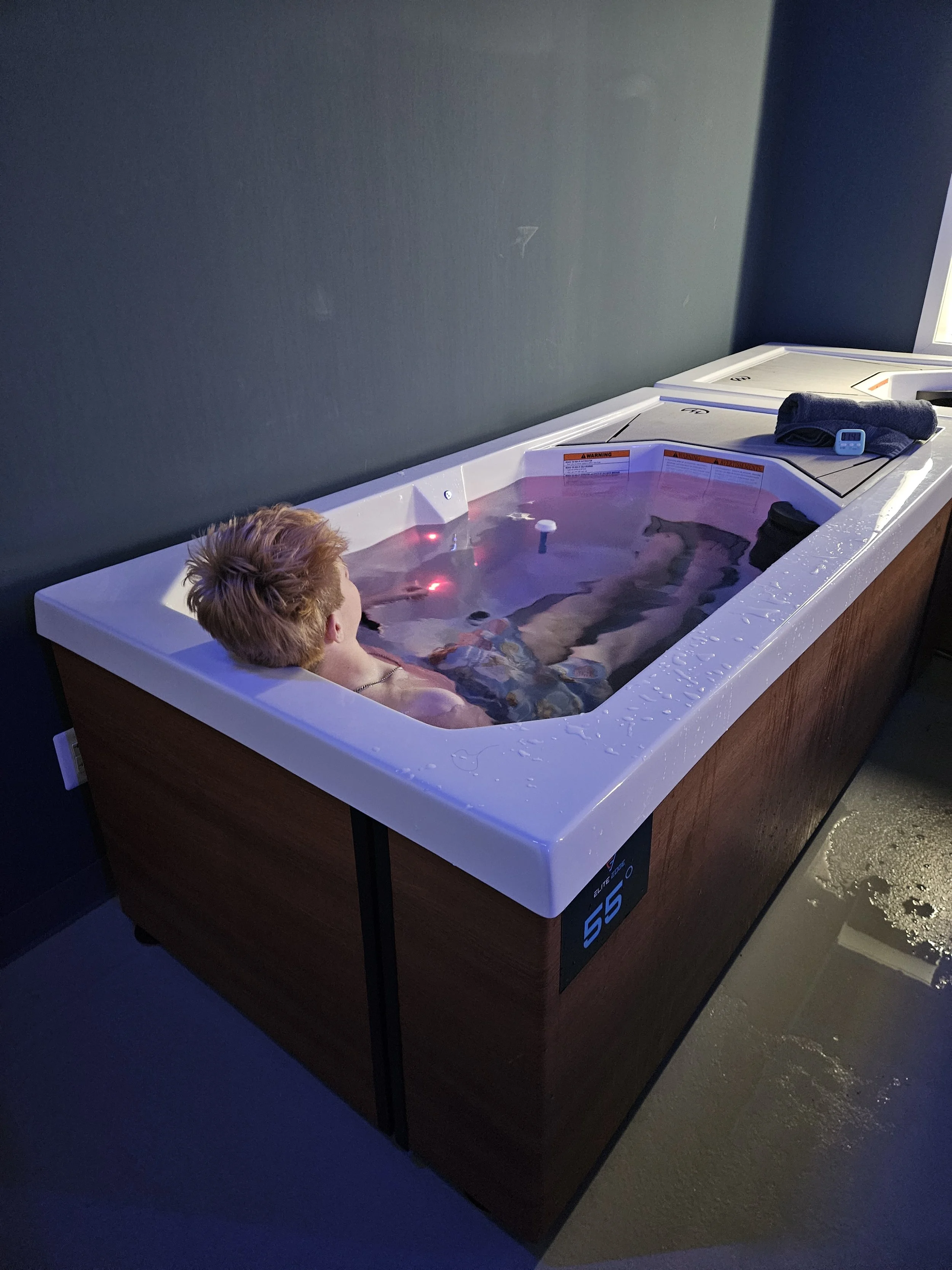 Person relaxing in a hot tub with water, towels, a digital thermometer, and a towel on the edge, against a dark wall.