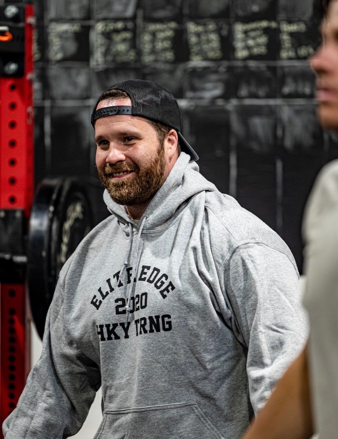 A man with a beard wearing a black baseball cap backwards and a gray hoodie with black lettering, smiling indoors.