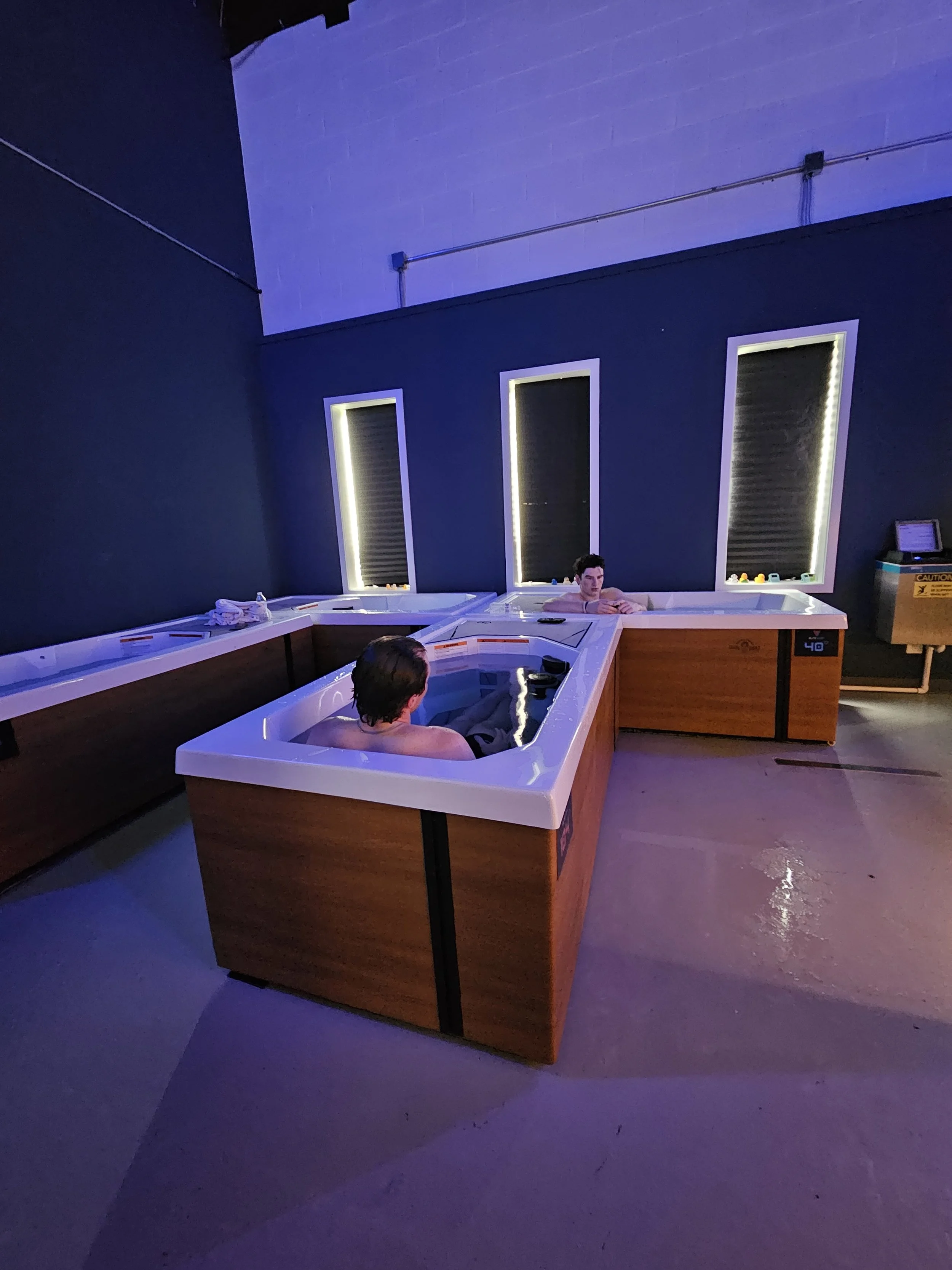 Two people in hot tubs inside a dark, modern room with blue lighting and three tall, narrow windows with black blinds