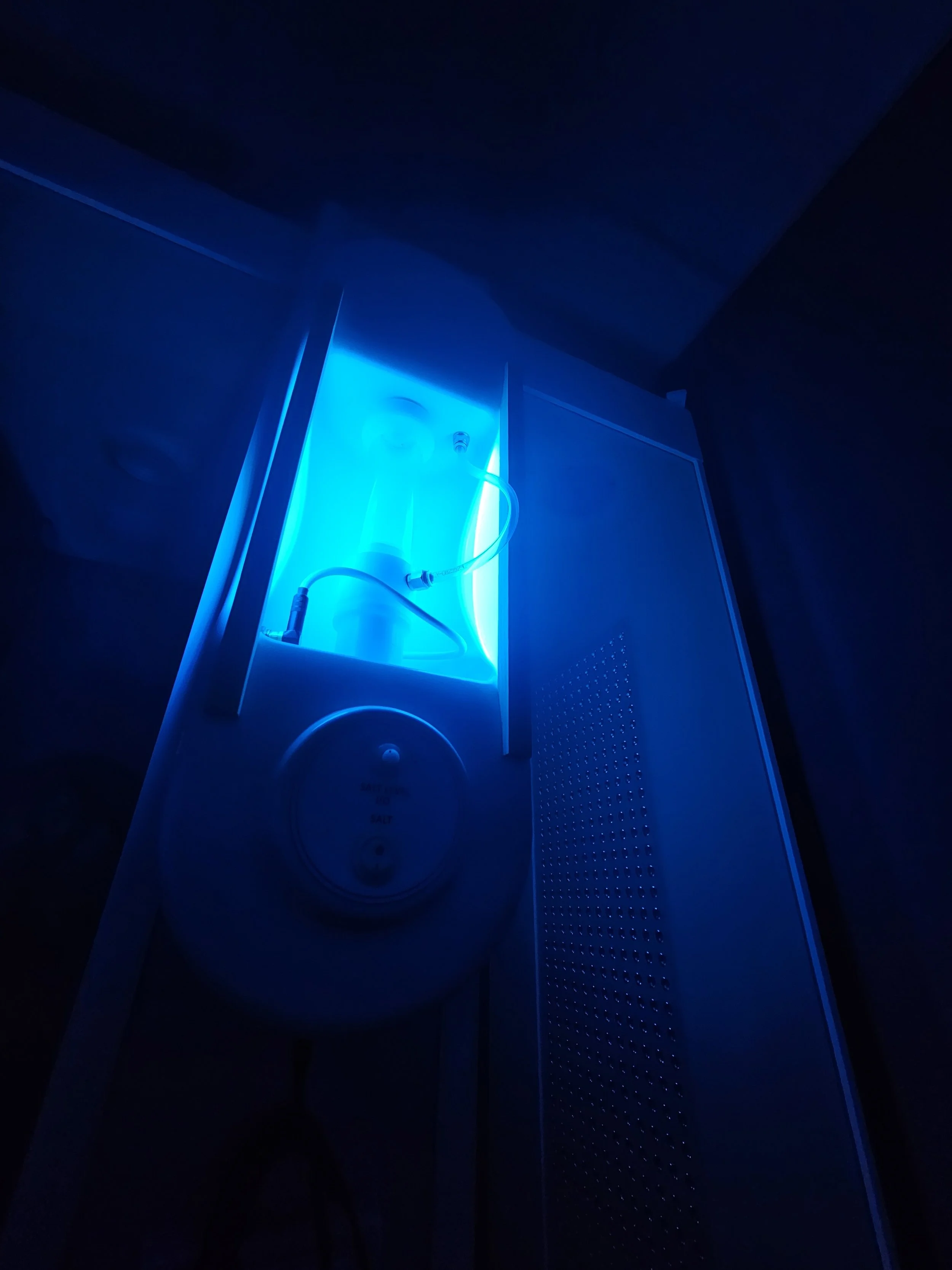 A dimly lit room with blue lighting from a neon sign. The image shows a control panel or device with a round dial and a small screen, mounted on a wall next to a perforated metal surface.