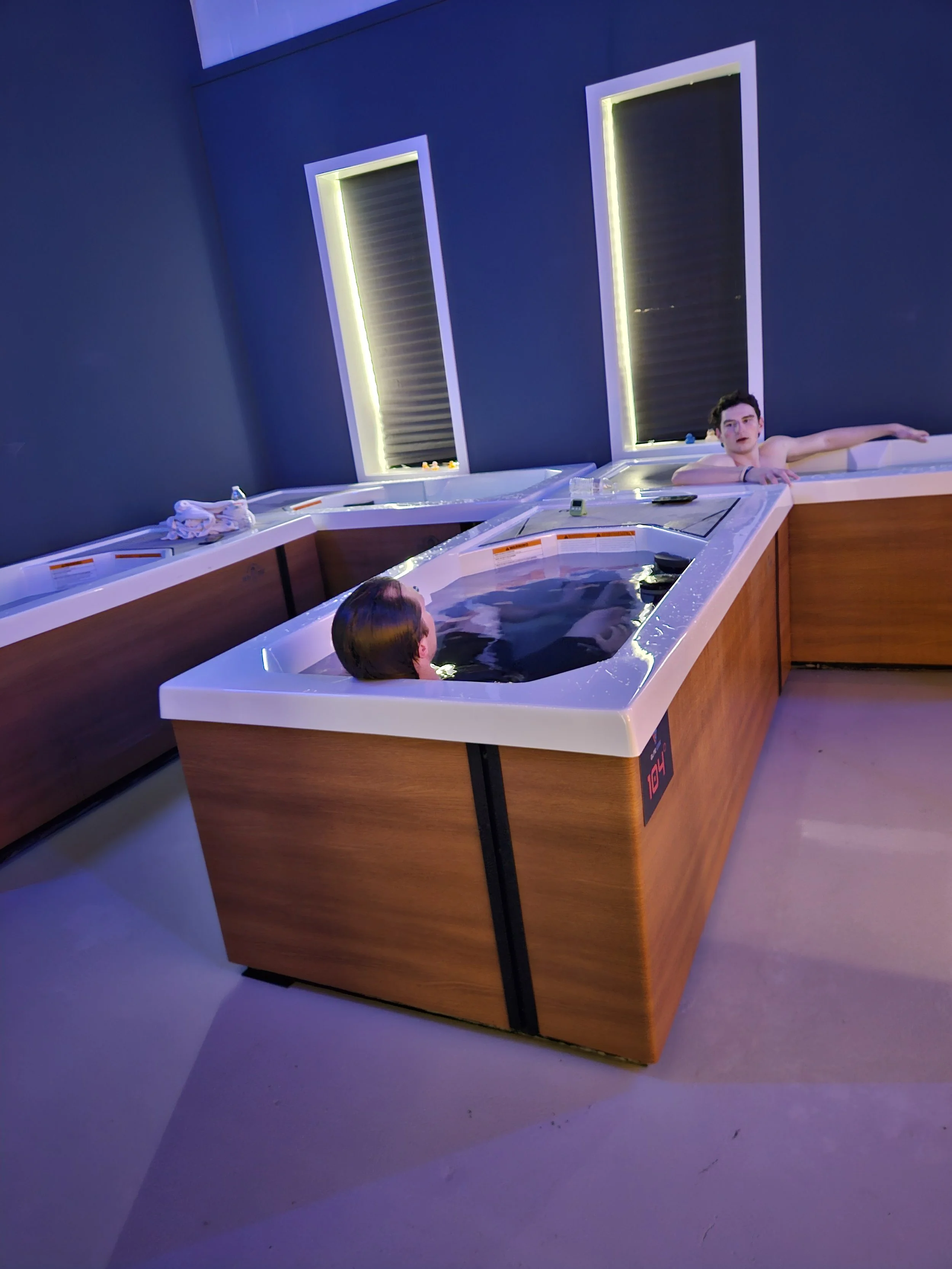 Two people relaxing in a hot tub in a modern spa room with dark blue walls and vertical lighted windows.