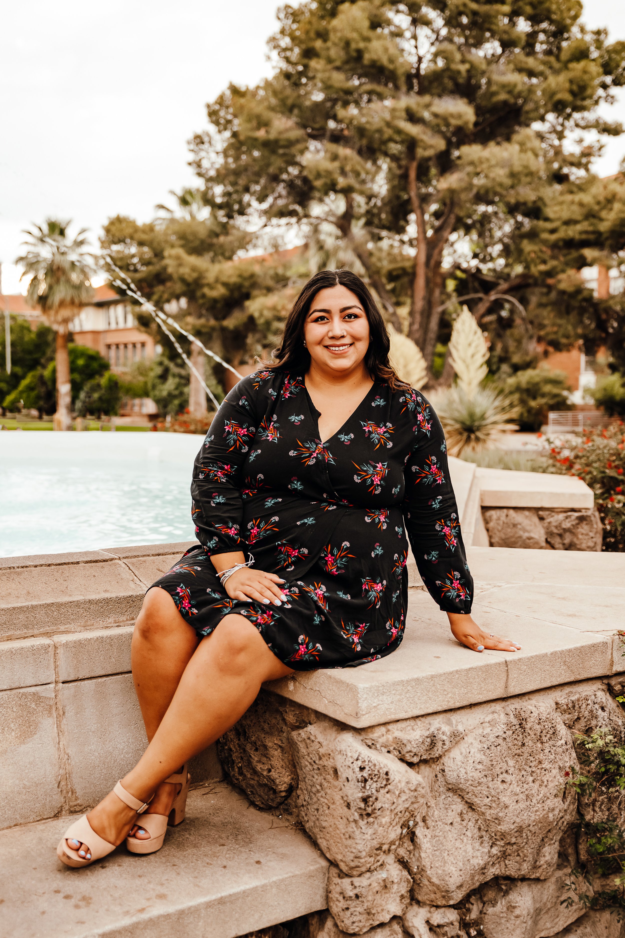 A woman in a black floral dress smiling and sitting on a stone ledge near a fountain in an outdoor park setting with trees and buildings in the background.