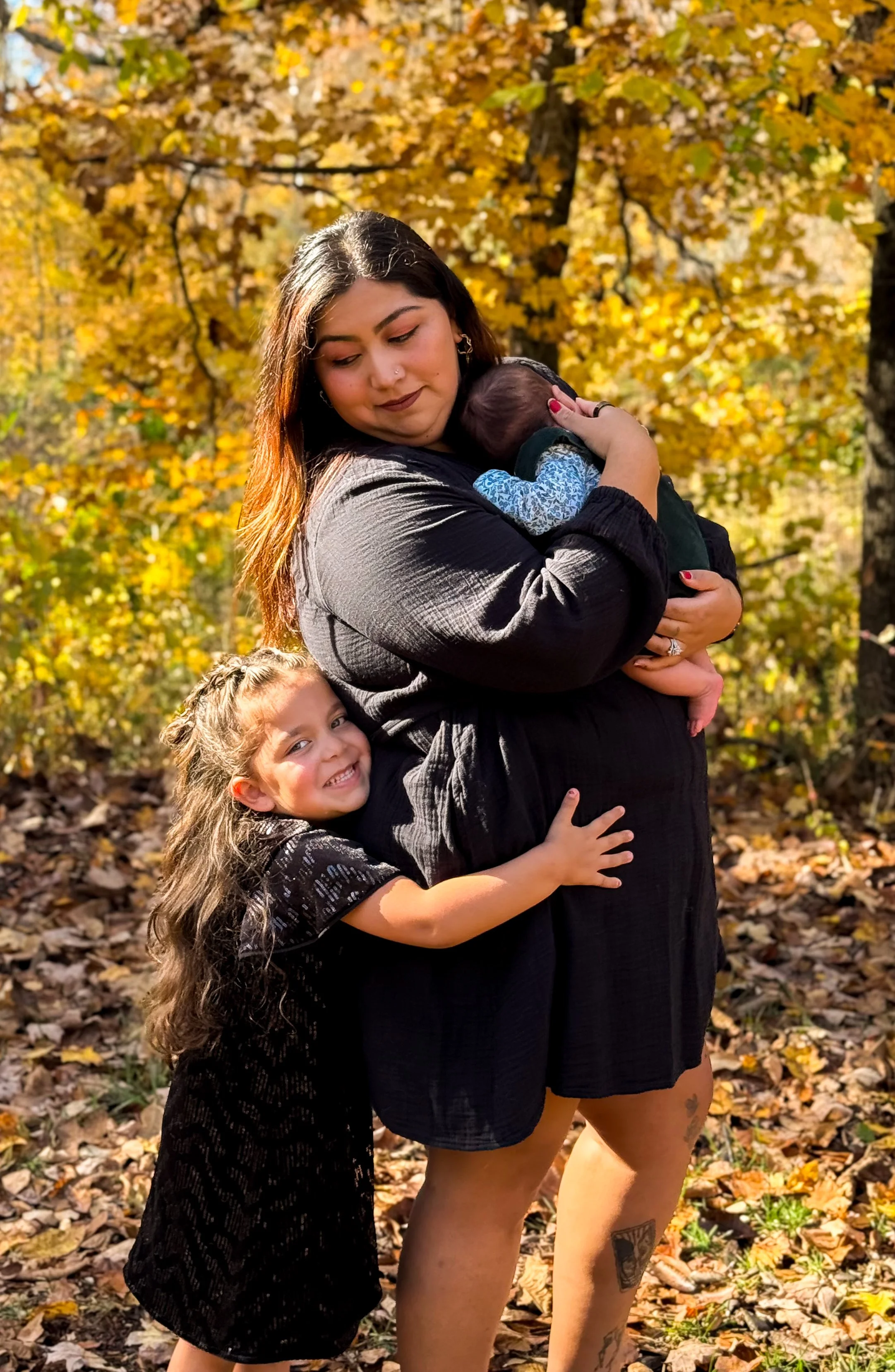 A woman holding a baby in a forest with fall foliage, and a young girl hugging her leg, all smiling and enjoying the autumn day.