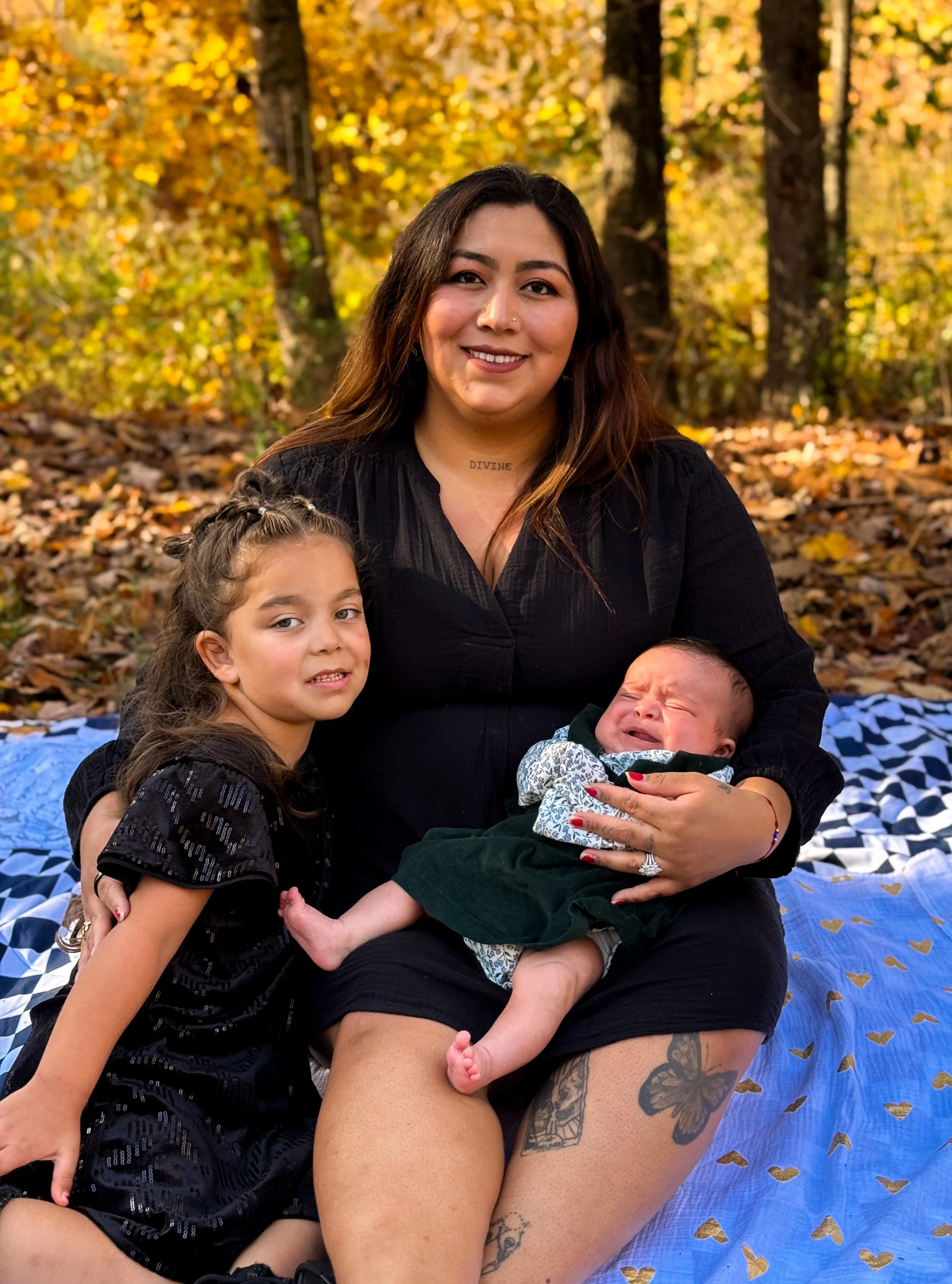 A woman with long dark hair sitting outdoors on a blanket, holding a crying baby in her lap, with a young girl kneeling beside her, in a park during autumn with yellow and orange leaves on the trees and ground.