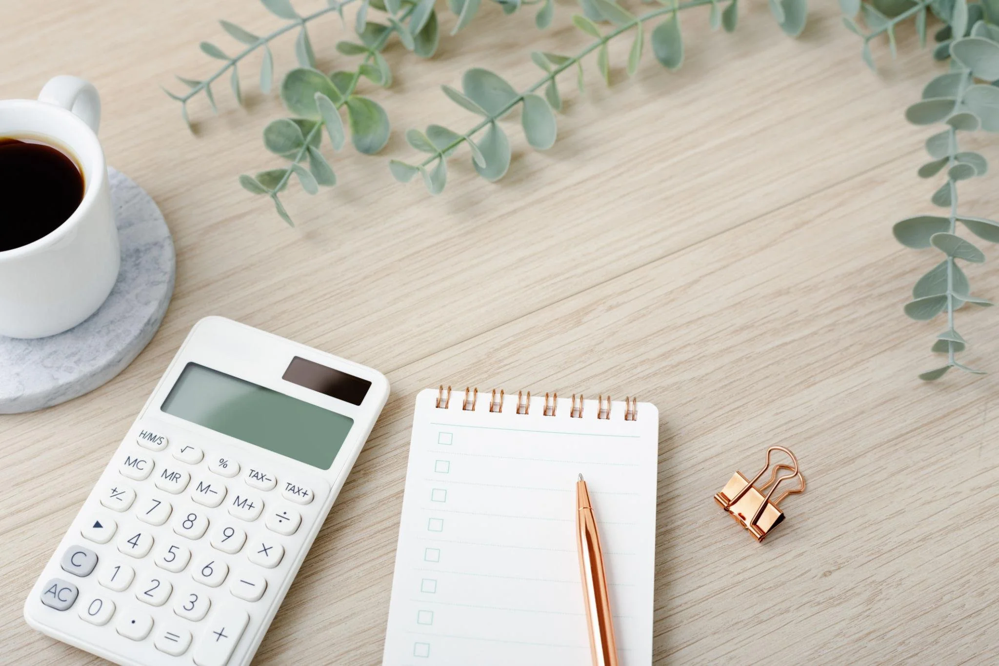 A wooden desk with a white calculator, a small notepad with a rose gold pen, a copper binder clip, a white mug of coffee on a gray coaster, and green eucalyptus leaves.
