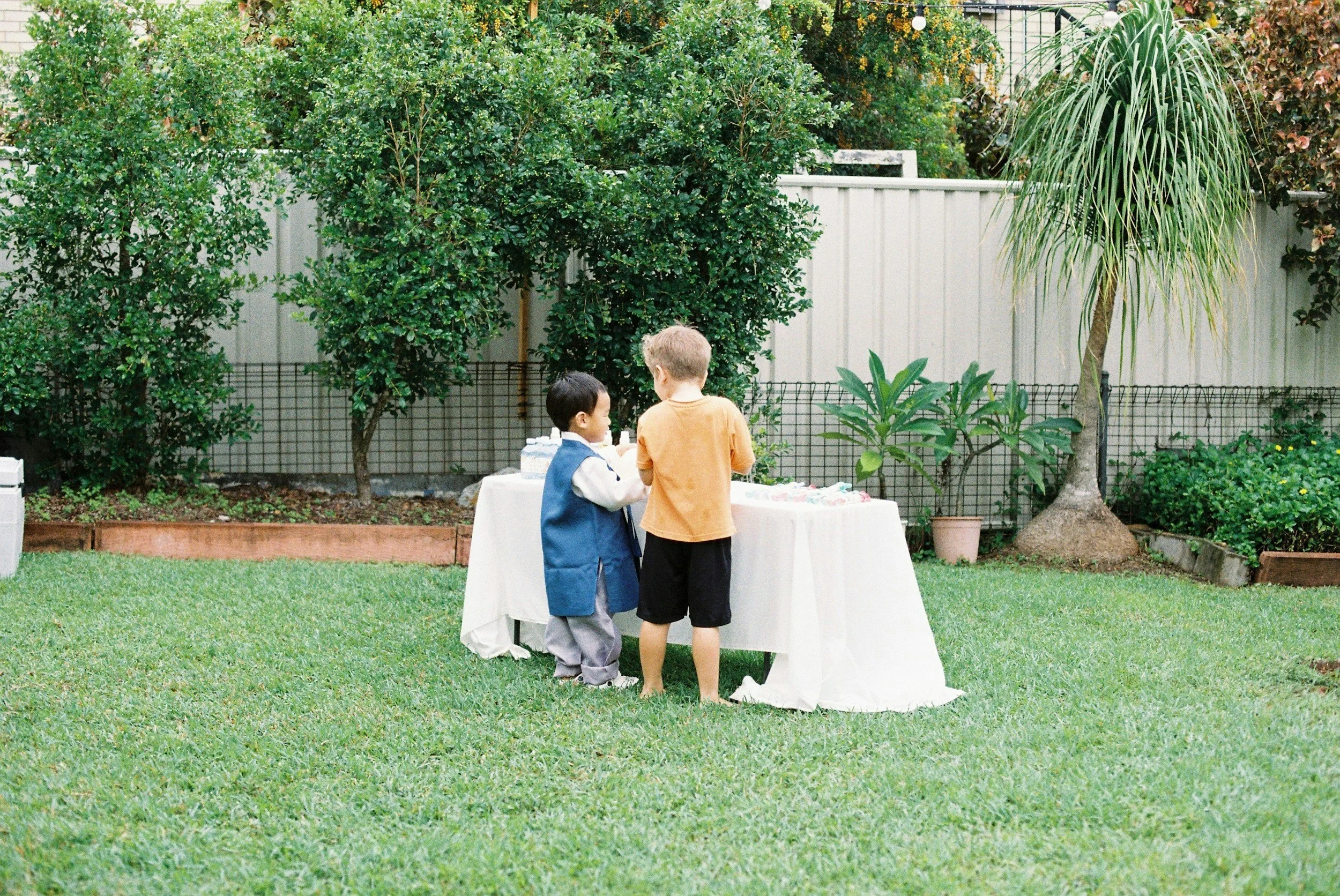 Zwei Jungen stehen an einem weißen Tisch im Garten, während sie miteinander sprechen, im Hintergrund grüne Büsche und Bäume.