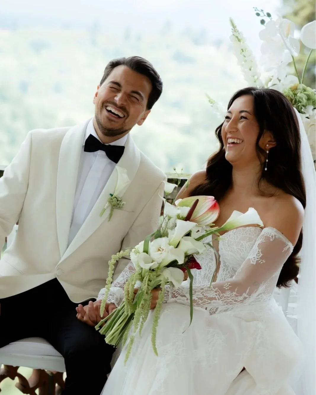 Ein lachendes Brautpaar bei einer Hochzeit, der Bräutigam trägt einen weißen Anzug mit schwarzer Fliege, die Braut trägt ein weißes Hochzeitskleid und hält einen Blumenstrauß.