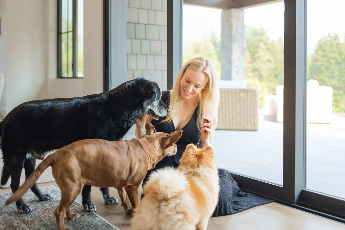 A woman sitting on the floor near a glass door, smiling and interacting with three dogs of different breeds and sizes inside a home.
