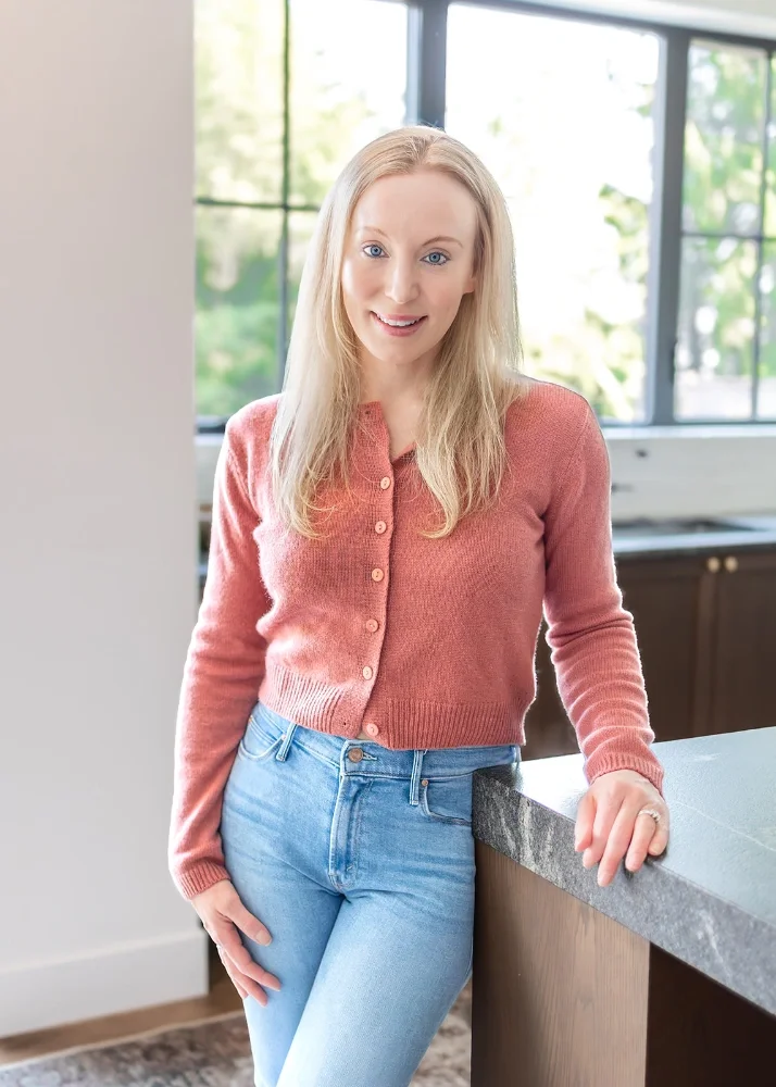Therapist Britta Jensen wearing a pink cardigan and blue jeans, leaning on a kitchen counter in a bright room with large windows showing greenery outside.