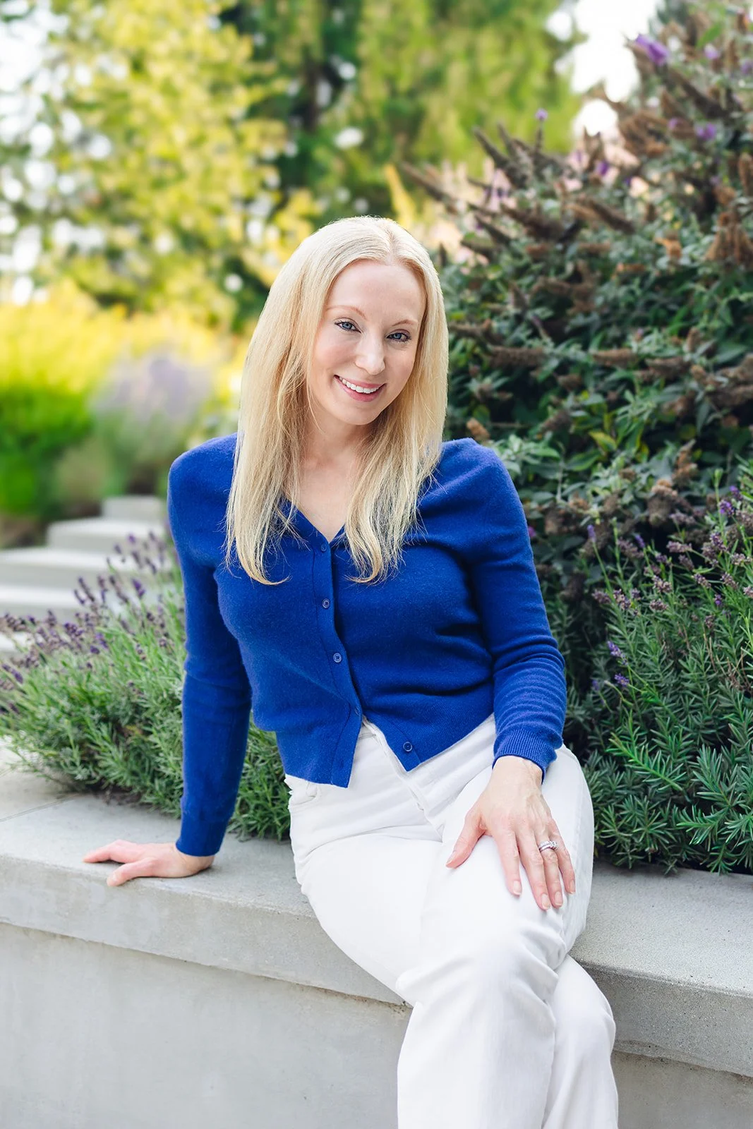 A woman with long blonde hair, wearing a blue cardigan and white pants, sitting on a concrete edge in a garden with green trees and purple flowers.