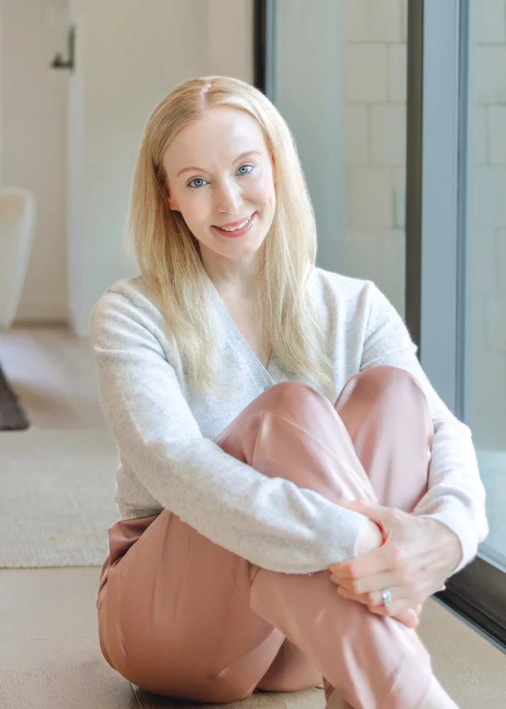 Therapist Britta Jensen wearing a light gray sweater and pink pants, sitting on the floor by a large window, smiling at the camera.