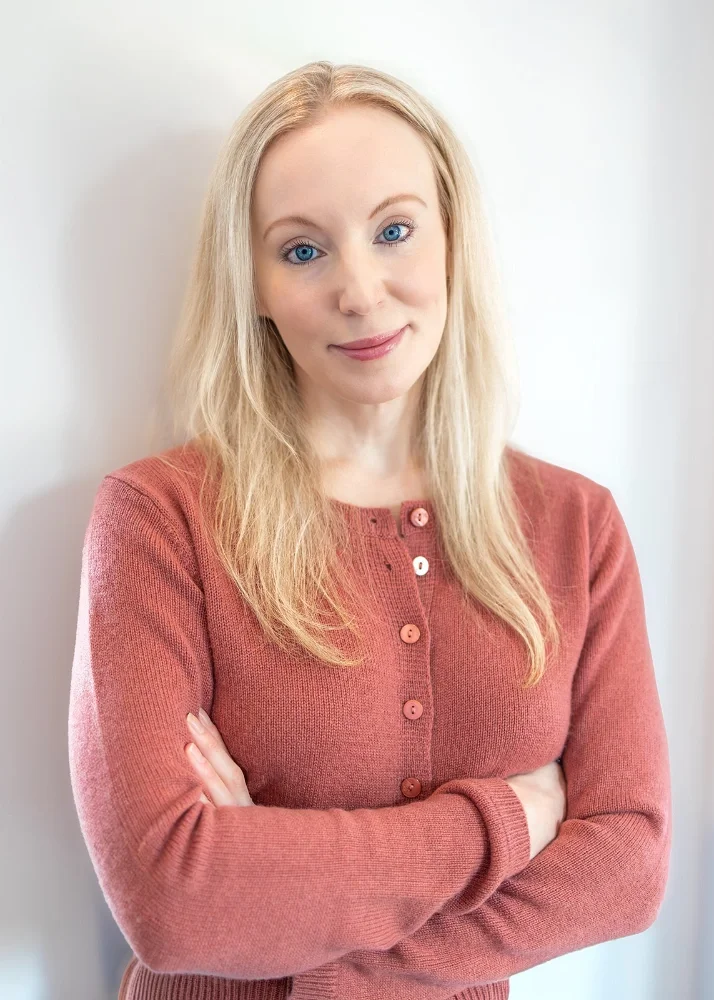 A woman with blonde hair and blue eyes, wearing a pinkish-red sweater, standing with her arms crossed and smiling softly against a plain, light-colored background.