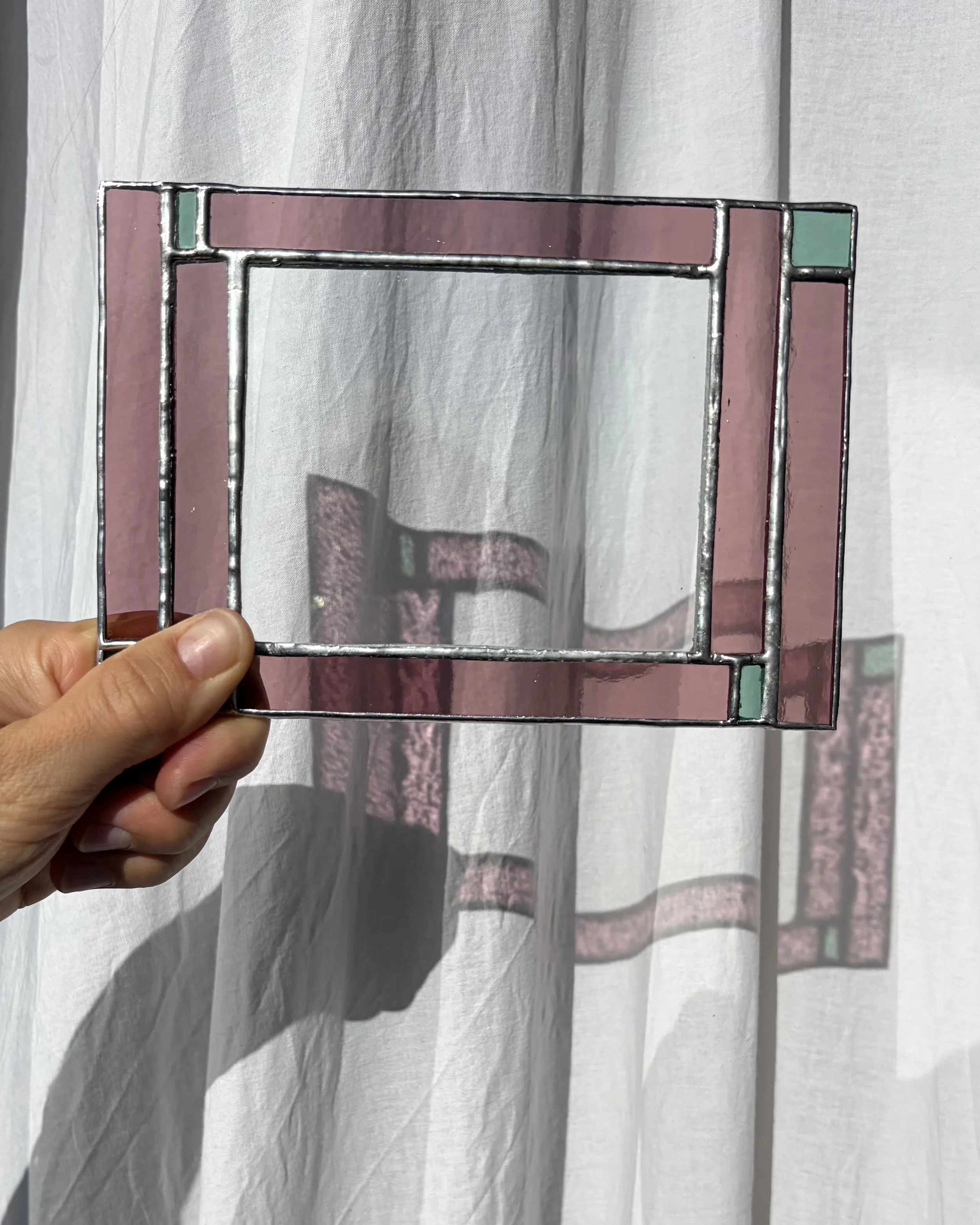 Hand houdt een decoratief metalen lijst met roze, groene en doorzichtige glas-in-lood stukken, tegen een witte, licht doorlatende gordijn achtergrond.
