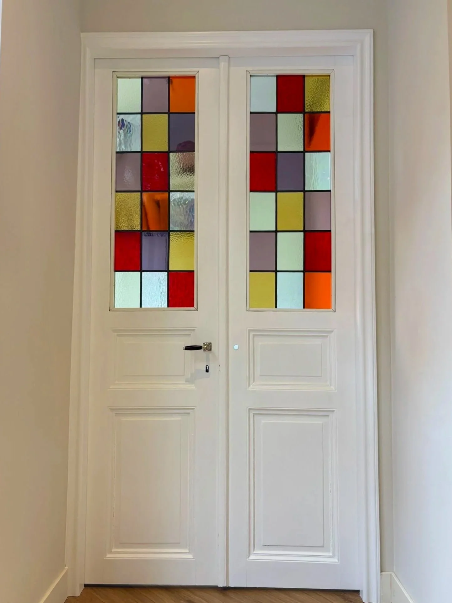 Een witte deur met vier gekleurde glazen panelen, twee op elke vleugel, met een zwarte deurknop en een sleutel. De panelen bevatten rode, gele, paarse, oranje, en groene gekleurde glazen stukken.