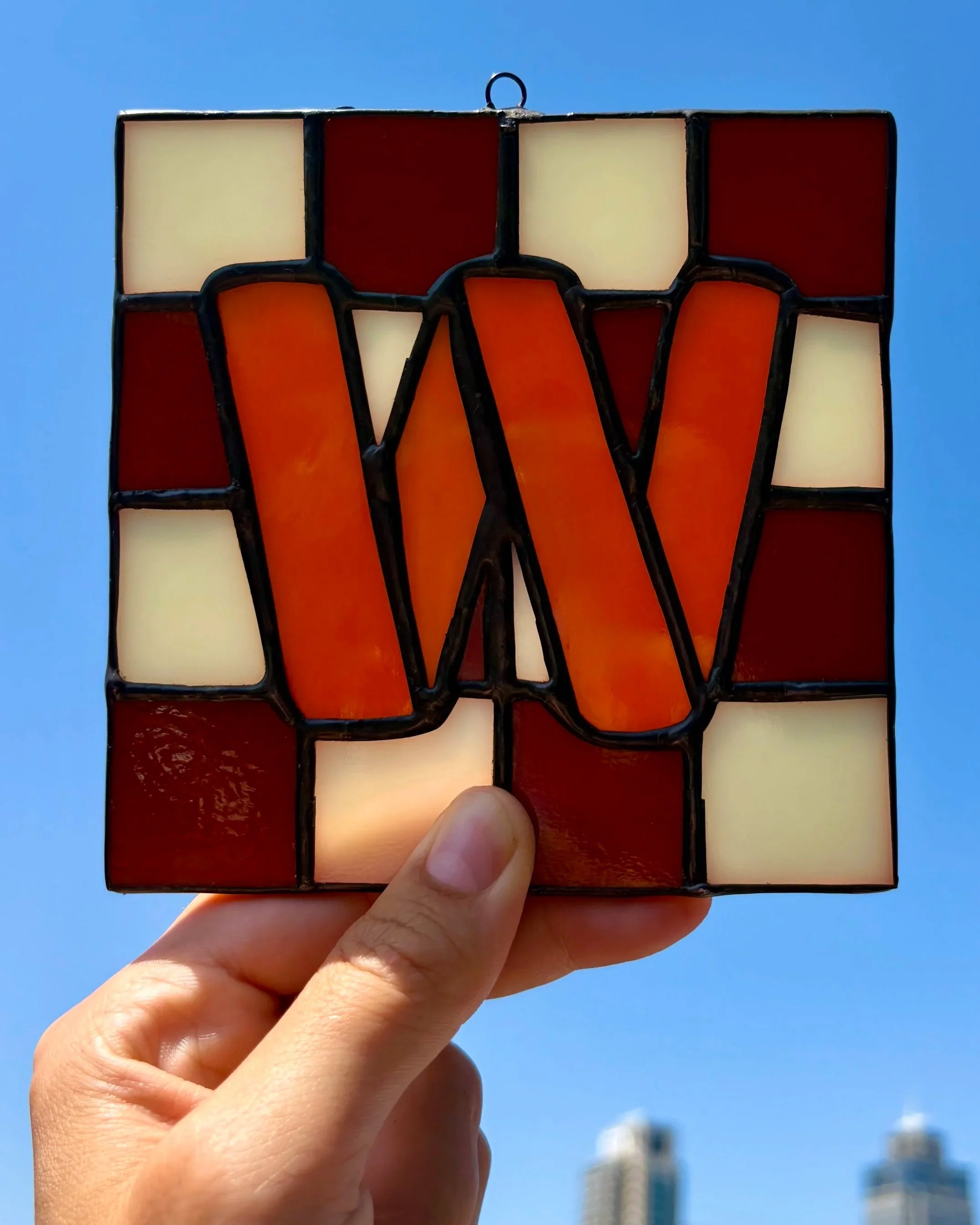 Een hand houdt een geblokt tegeltje van glas in lood vast, met de letter W, tegen een blauwe hemel.