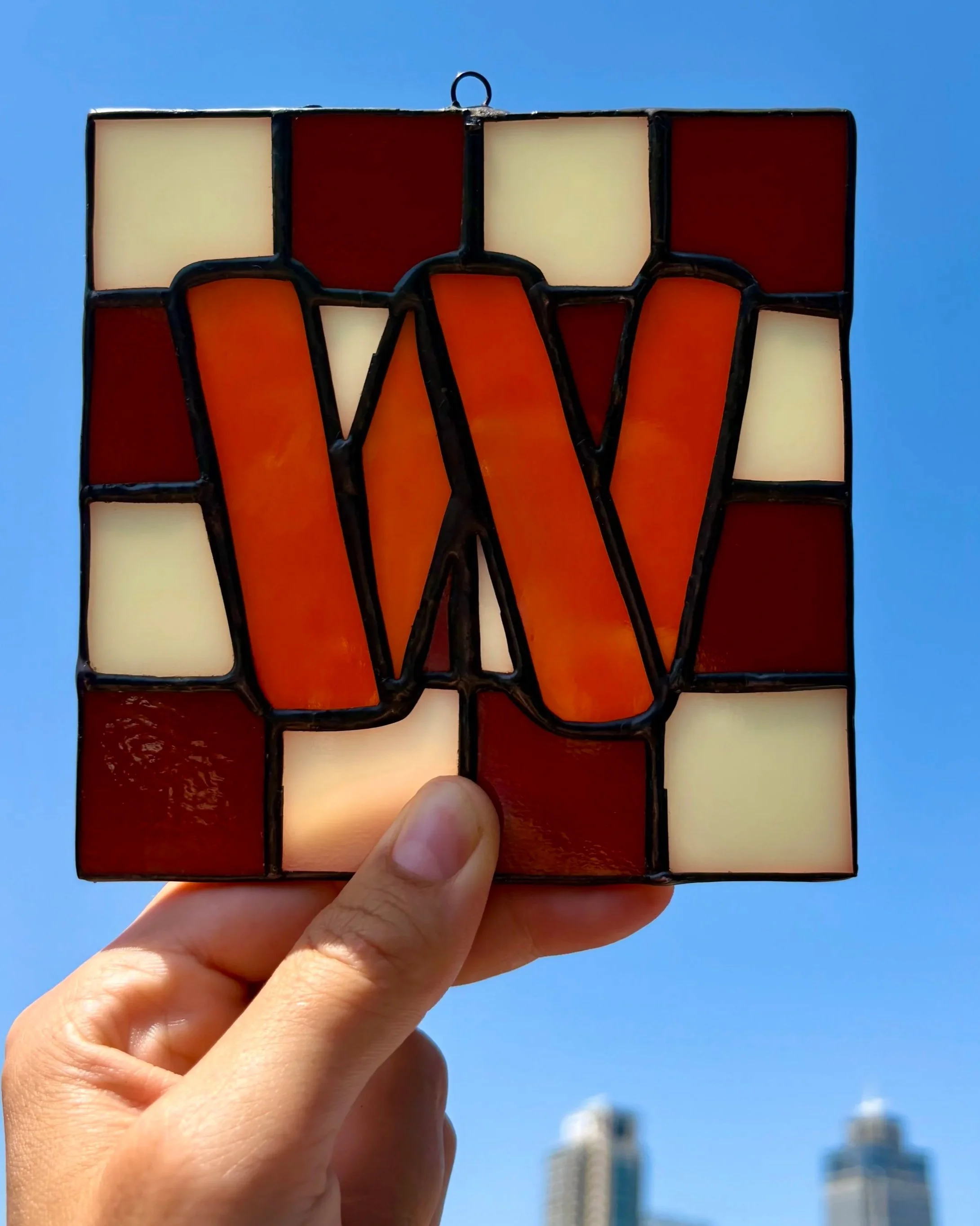 Een hand houdt een glazen kunstwerk vast dat uit gekleurde stukken bestaat, met een blauwe lucht en een paar hoge gebouwen op de achtergrond.