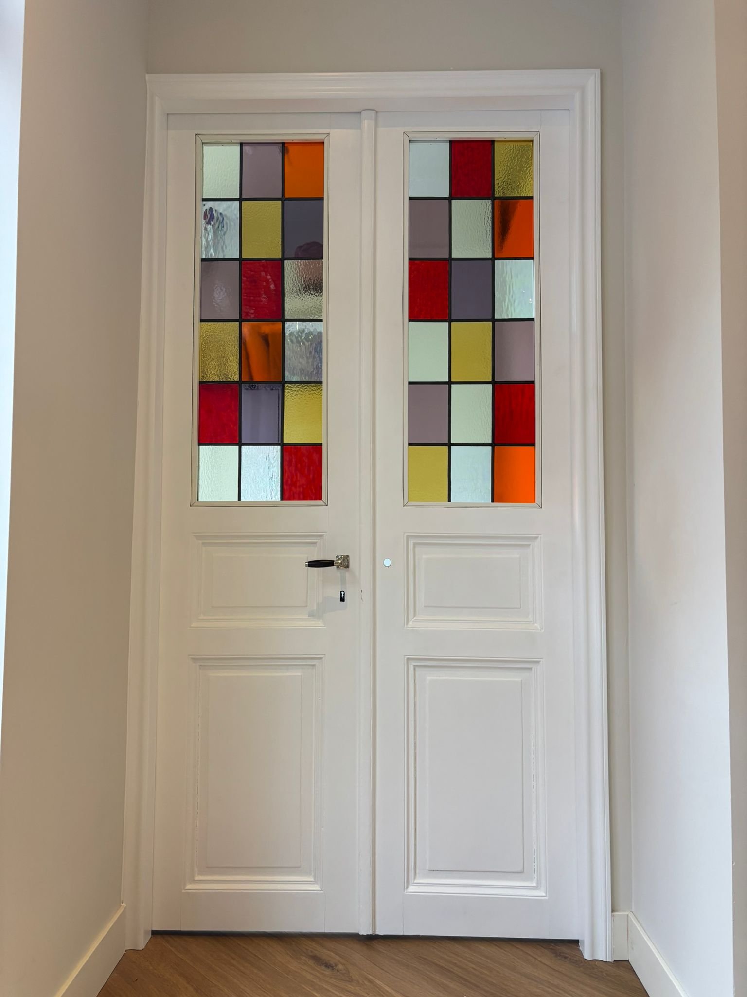 Dubbele witte deur met gekleurde glas-in-lood panelen, omgeven door witte deurkozijn en grijze muur.
