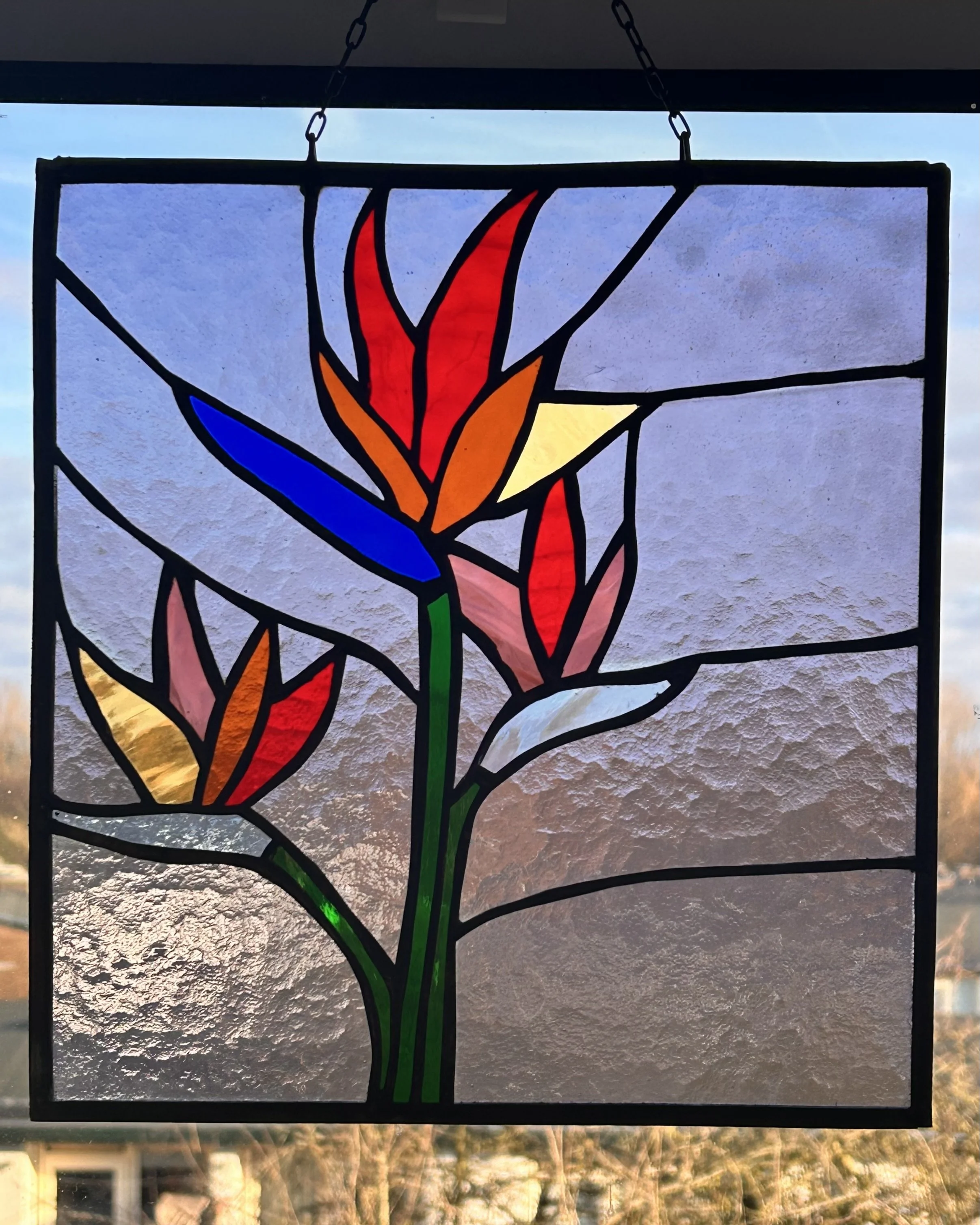 Een gekleurde glas-in-lood raampaneel met een abstract ontwerp van bloemen en bladeren, met een achtergrond van een blauwe lucht en een landschap.