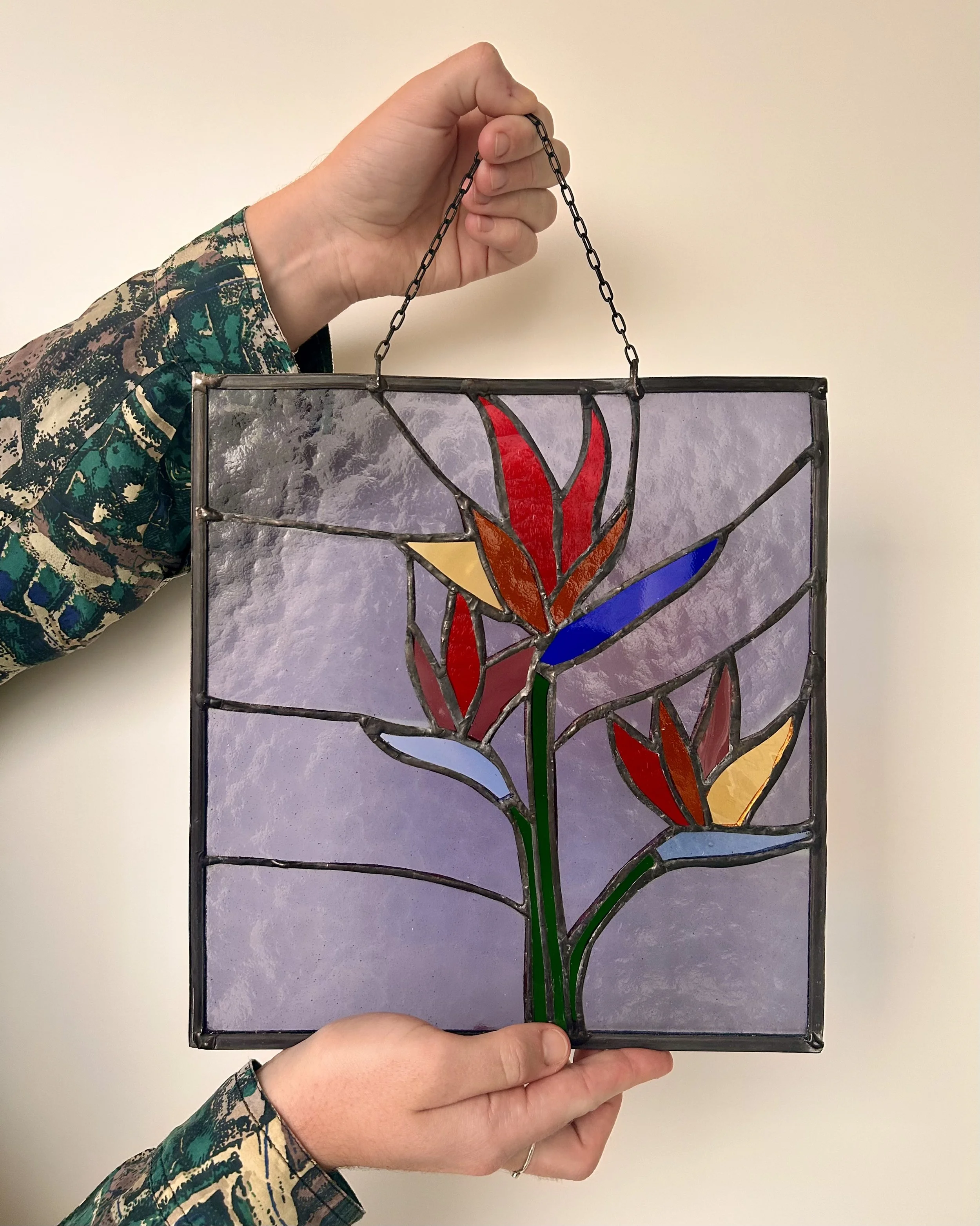Persoon houdt een glas in lood raam vast met een bloemdesign in meerdere kleuren, zoals rood, geel, blauw, en groen, tegen een witte muur.