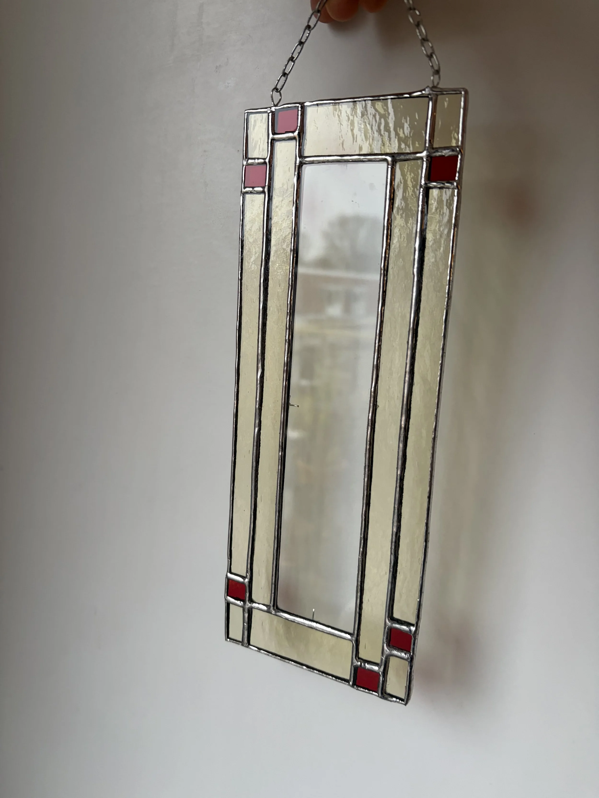 Een decoratief metalen en gekleurde spiegel met een modern, artistiek ontwerp. Hangt aan een ketting tegen een effen muur.