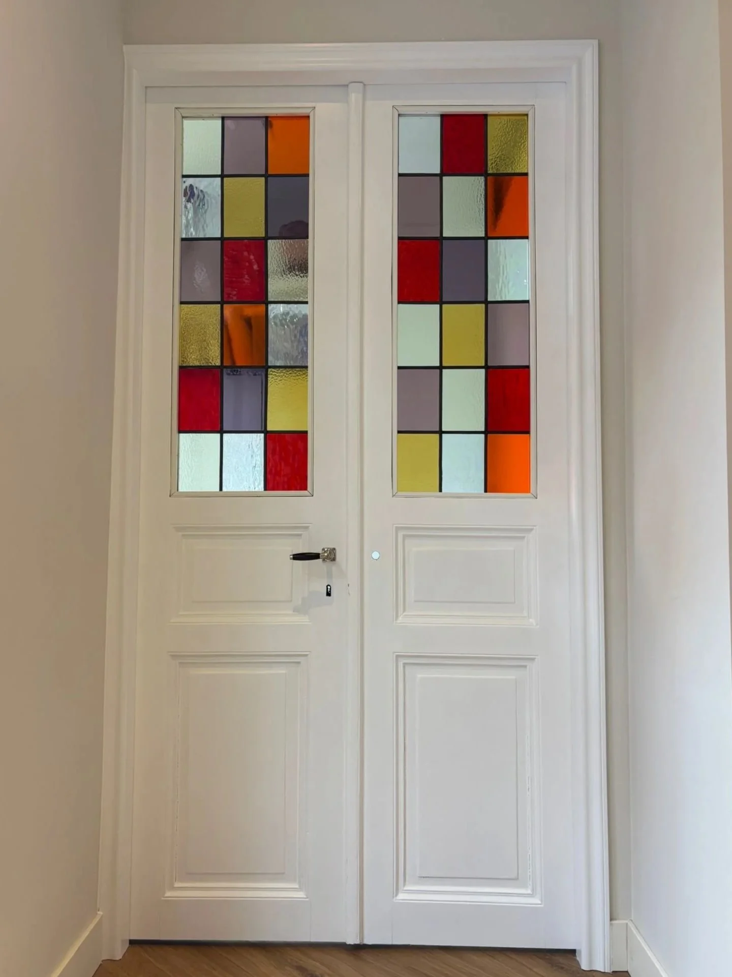 Witte houten deuren met gekleurde glazen panelen, omringd door een witte muur.