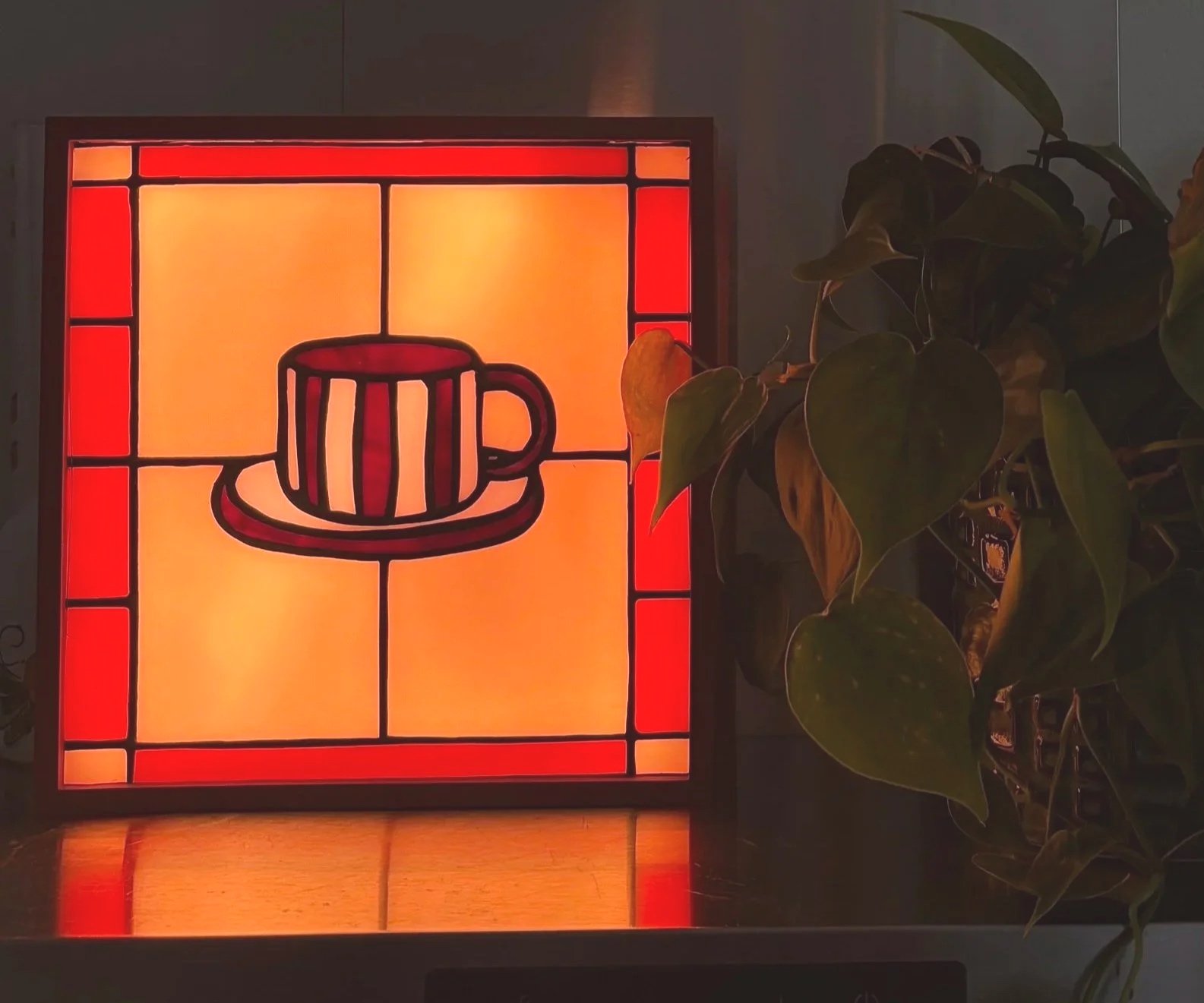 Een kleurrijk gestileerd glas-in-lood raam met een kopje koffie afgebeeld, naast een groene plant.