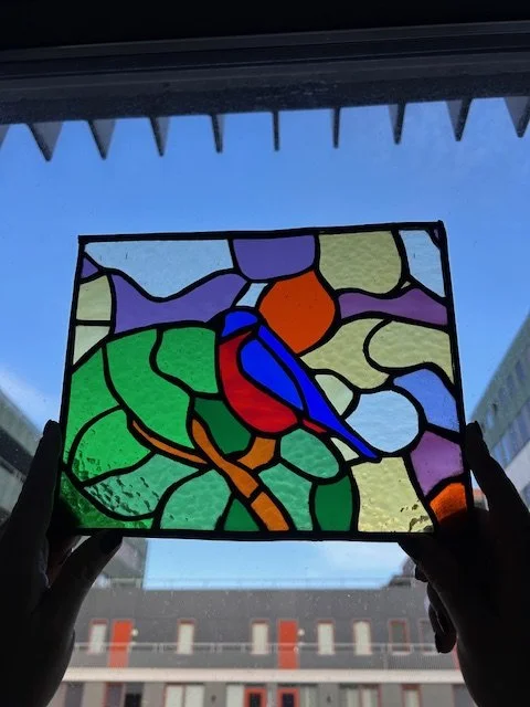 Een doorzichtige gekleurde glas-in-lood raampaneel met een landschap en een vogeltje.