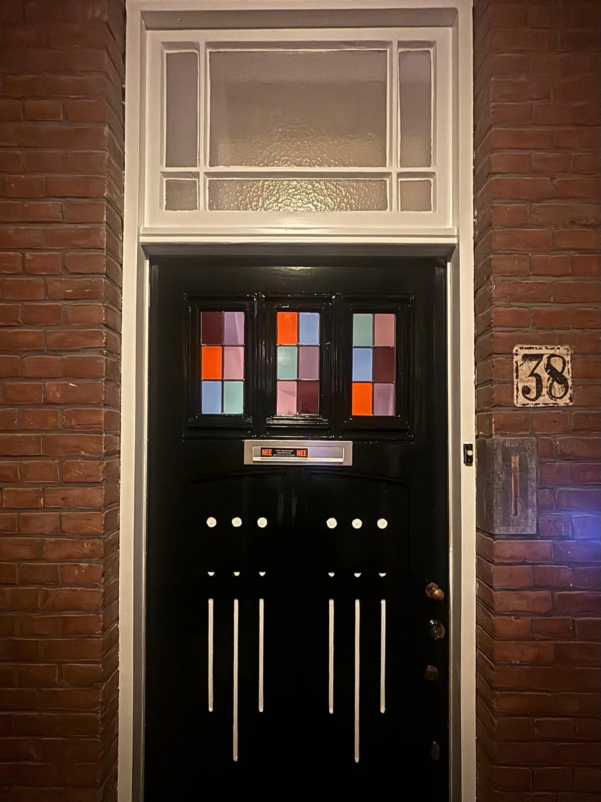 Deur met gekleurde glas-in-loodramen in een zwart omlijsting en een huisnummer 38 aan de muur, tussen bakstenen muren.