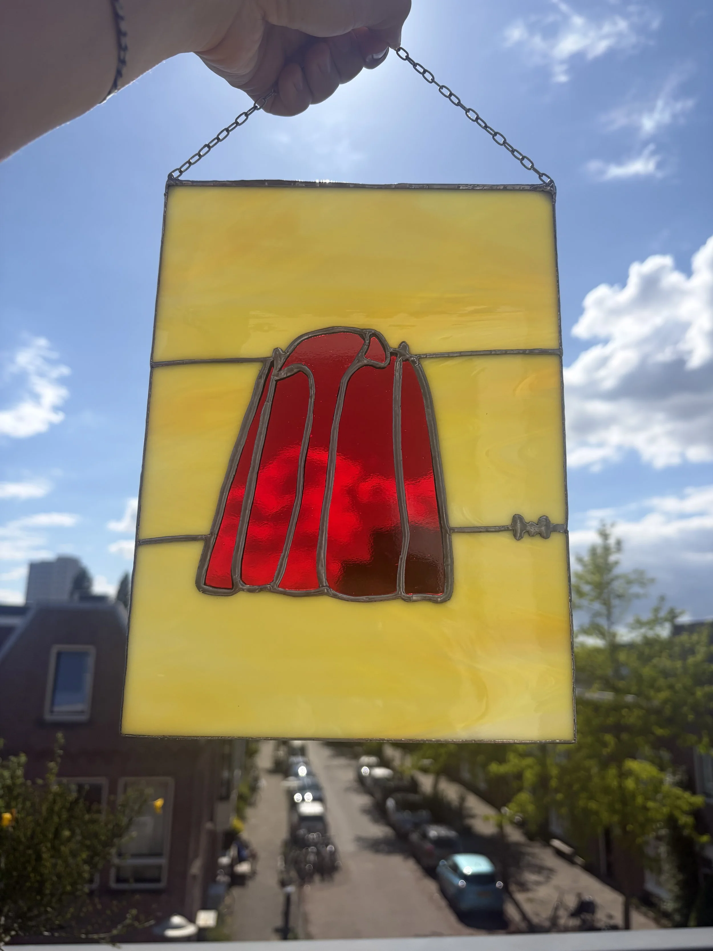 Een kleurrijk glas in lood kunstwerk met een rood jasje, dat wordt vastgehouden door een hand met een armband, tegen de achtergrond van een zonnige dag met blauwe lucht en enkele wolken.