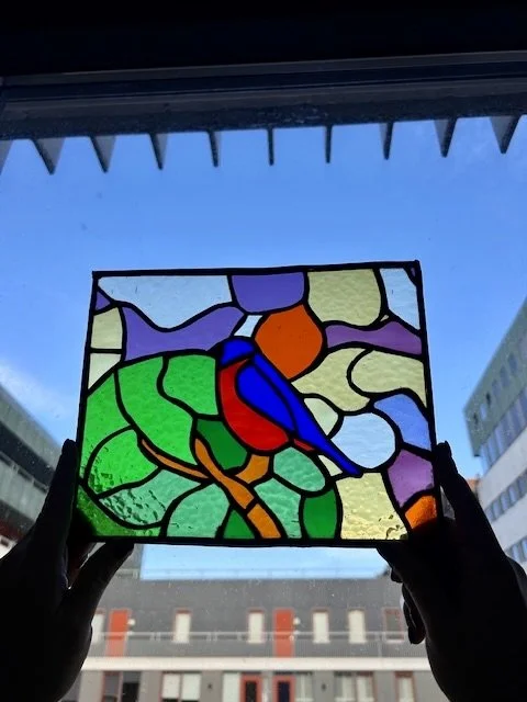 Violette, blauwe, groene, oranje en gele glas-in-loodraam in handen gehouden voor het raam met een stadsgezicht op de achtergrond.