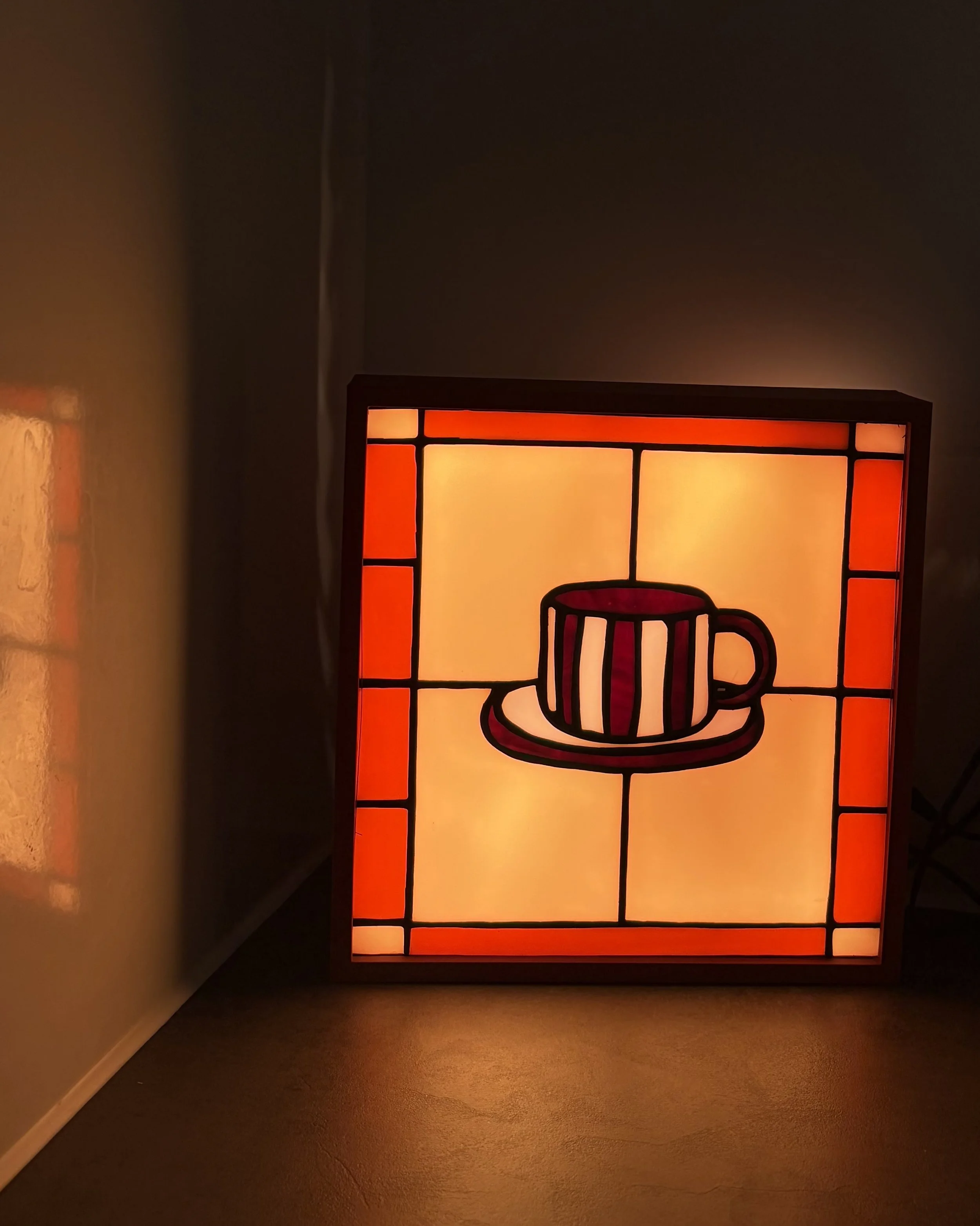 Een kleurrijk lamp met een abstract ontwerp van een koffiekopje met een rode rand en een handvat, geplaatst tegen een donkere muur, dat een warme gloed uitstraalt.