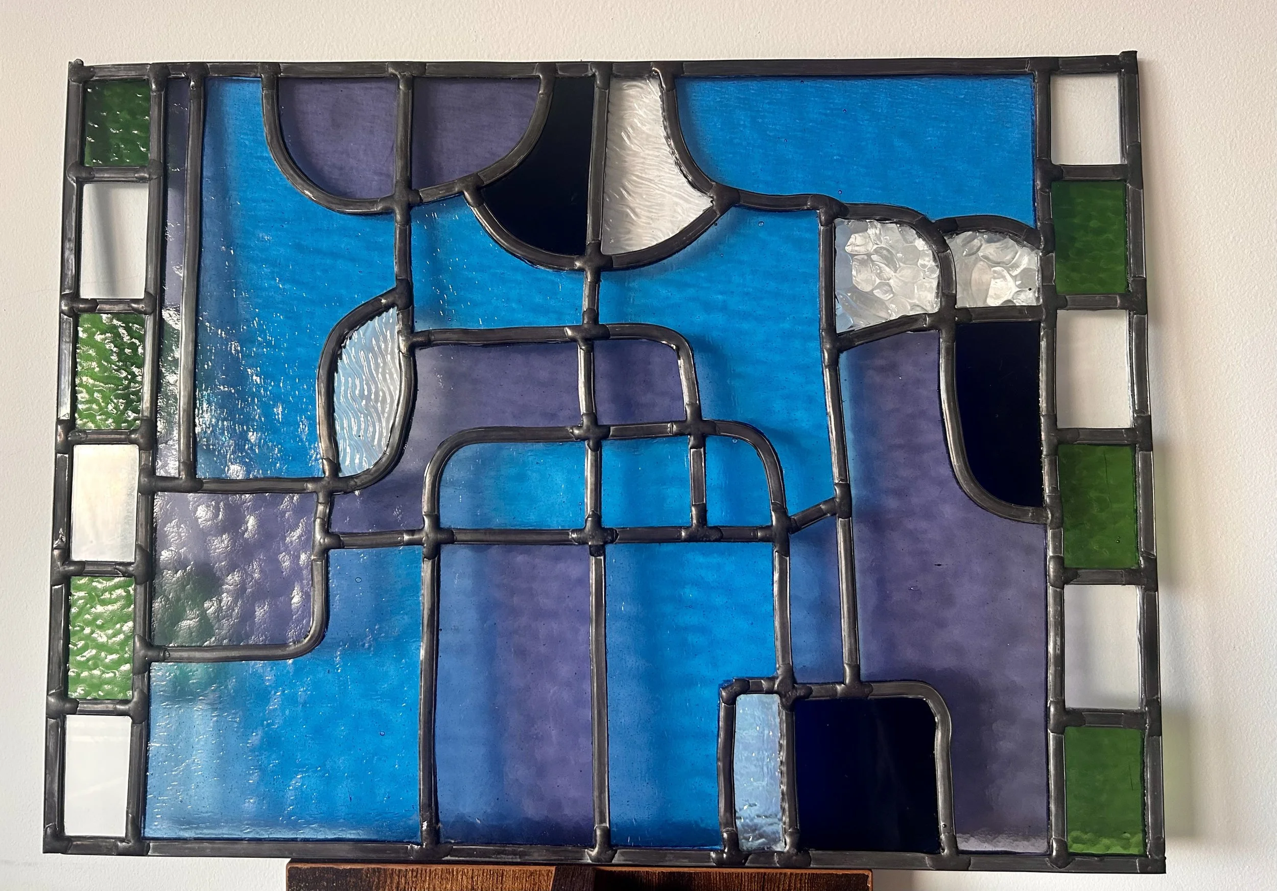 Een doorzichtige kunstzinnige glas-in-loodschildering met blauw, groen, paars, wit en zwart glas in een geometrisch patroon, bevestigd in een metalen frame.