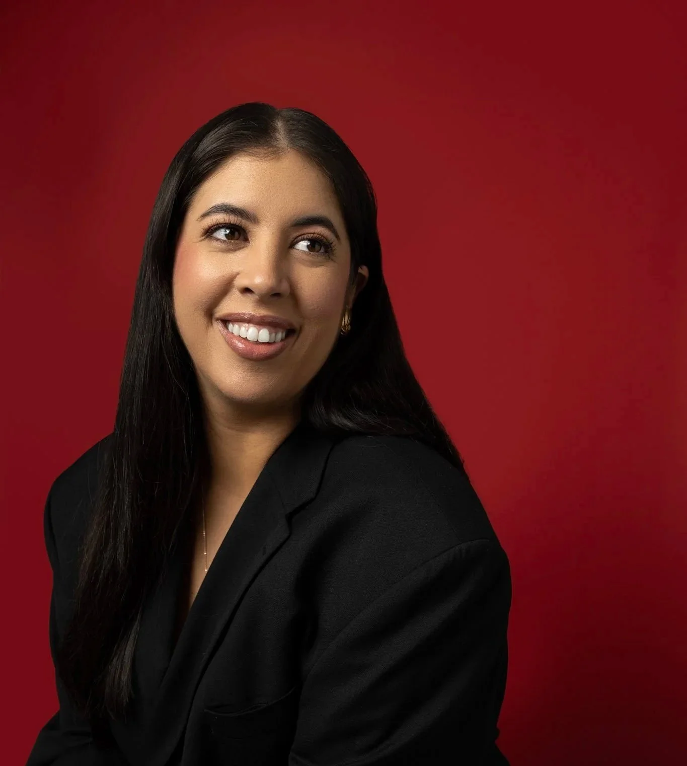 Professional headshot of author J. Mulberry. Jackie is looking away from the camera while smiling. She is wearing a black blazer in front of a red photo backdrop.
