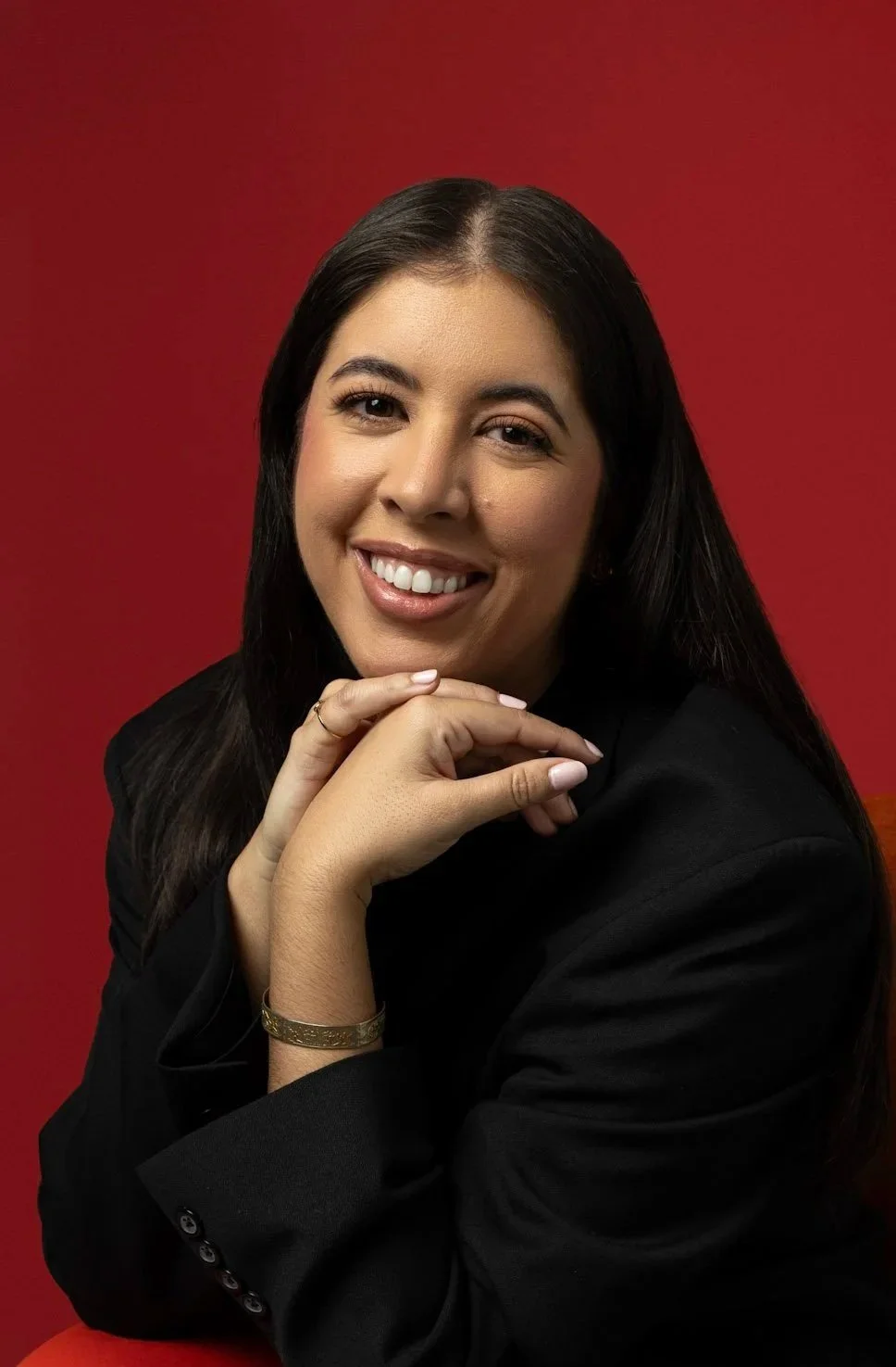 Professional headshot of author J. Mulberry. Jackie is resting her shin on her hands while smiling. She is wearing a black blazer in front of a red photo backdrop.