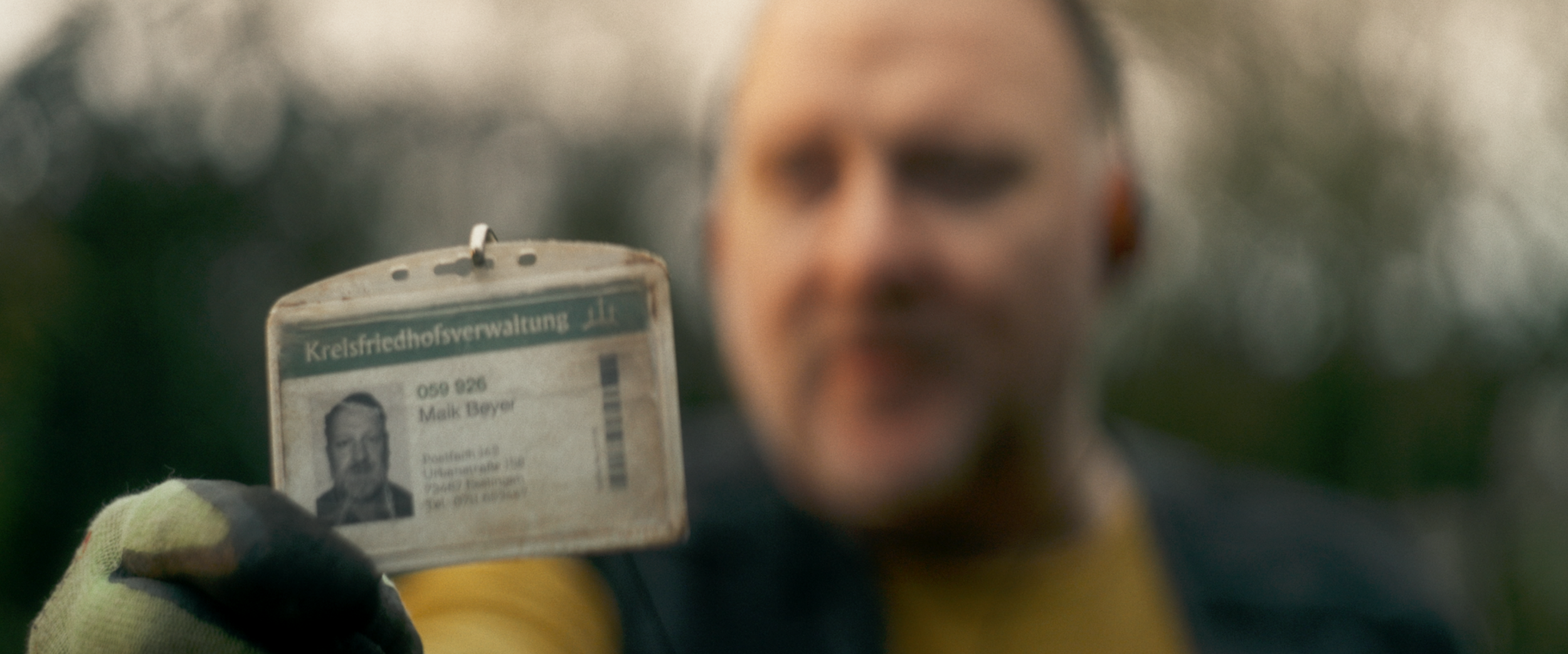 Ein Mann hält einen Ausweis der Kreishauptverwaltung mit einem Foto und Namen, während er in einem unscharfen Hintergrund steht.