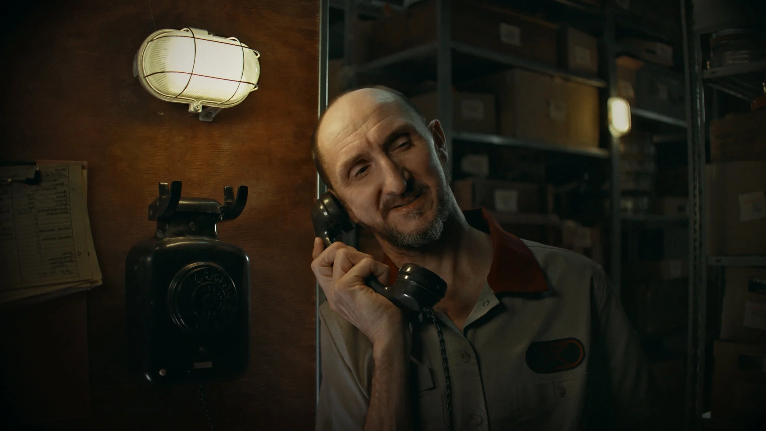 Ein Mann mit kurzen Haaren und Bart spricht an einem alten schwarzen Telefon in einem Lagerraum, im Hintergrund sind Regale und eine Lampe an der Wand.