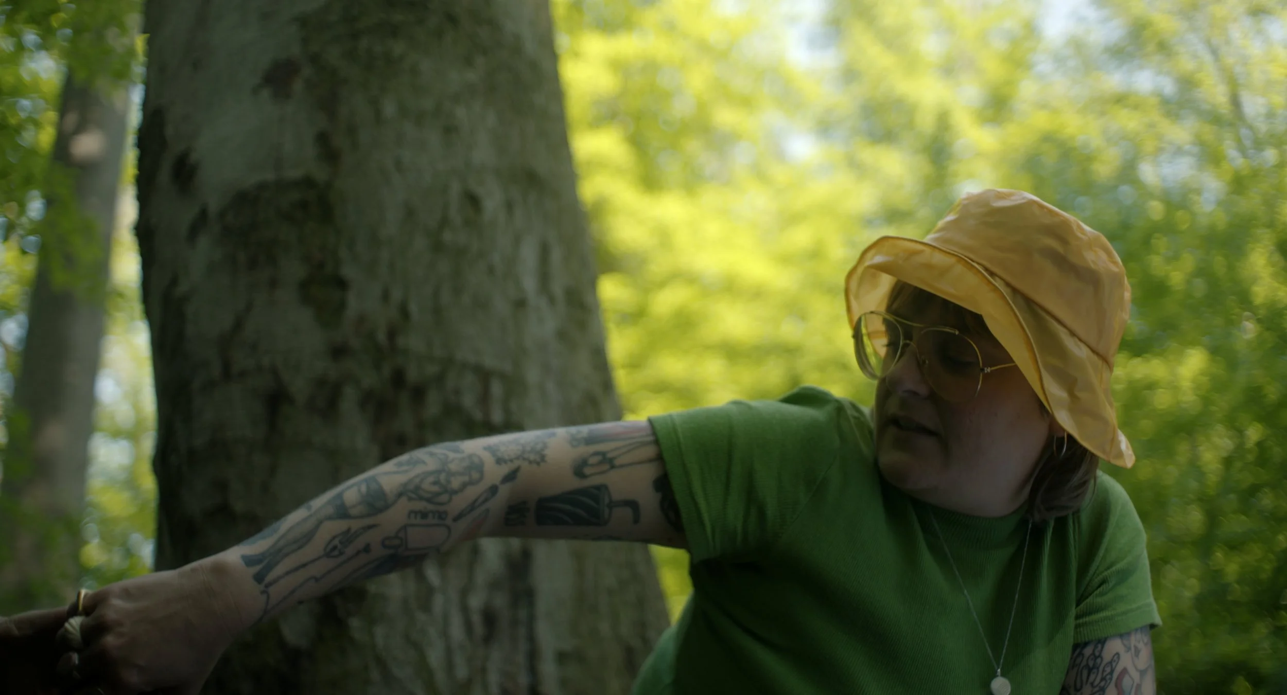 Eine Person mit Tattoos, Brille, gelber Hut und grüner Kleidung, die einen Baum im Wald berührt.