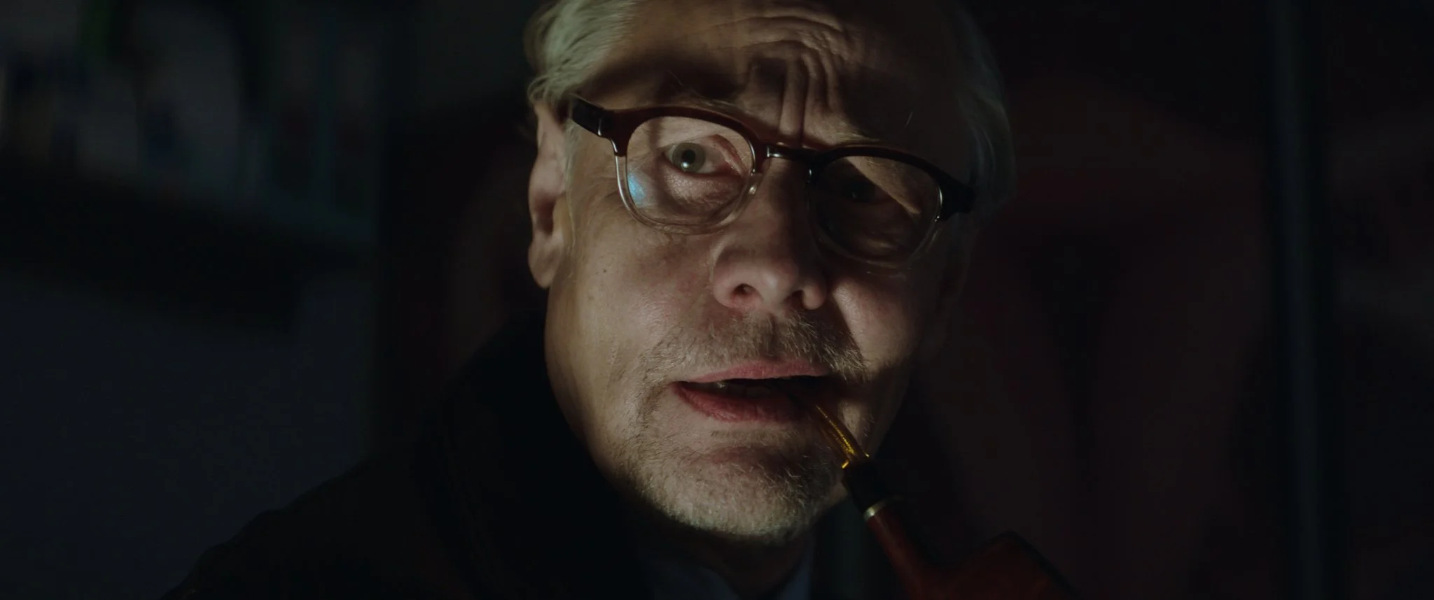 Ein älterer Mann mit Brille, grauem Haar und Schnurrbart raucht eine Pfeife in einem dunklen Raum.